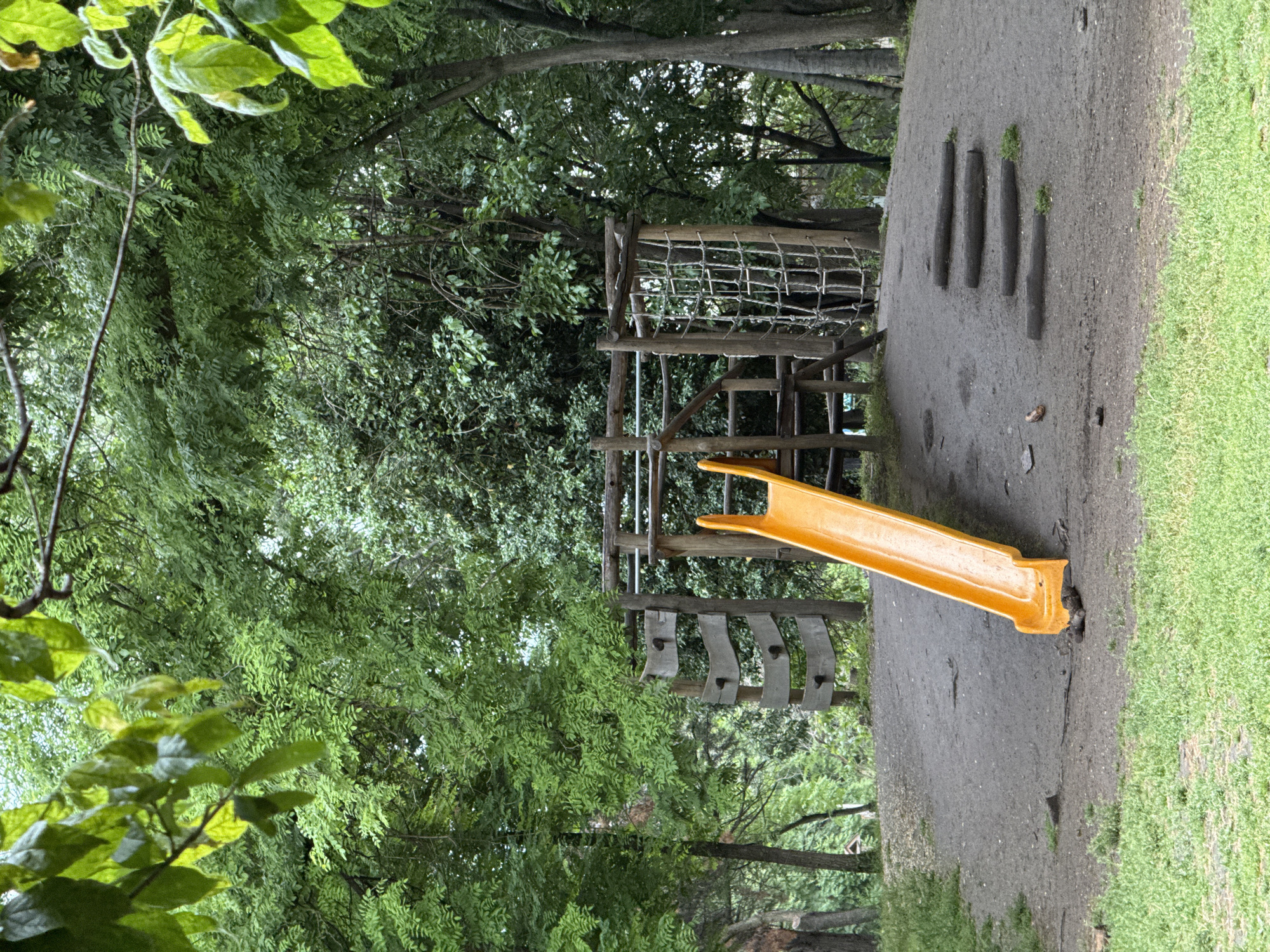 The image shows a children's playground in what appears to be a park setting. There is a wooden play structure with a yellow slide. The structure also includes a rope net climbing area and wooden steps. The ground is a mix of dirt and grass. The area is surrounded by trees. The image does not contain any people. The location is likely in Plovdiv, Bulgaria, though there are no explicit landmarks in the frame.