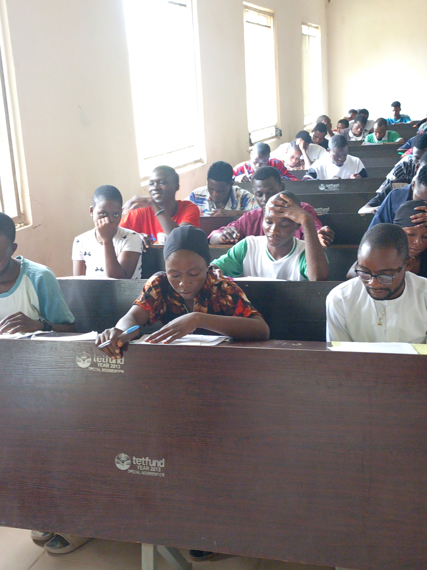 The image depicts a classroom scene in Akwanga, Nigeria. Students are seated at desks, most appearing to be taking an exam or completing a written assignment. The desks are labeled with "tetfund YEAR 2013 SPECIAL INTERVENTION." The students are of African descent, dressed in casual clothing. A man in a white shirt and glasses is seated in the front row, also writing. The classroom is lit by natural light from windows along the wall. The walls are painted in a light beige or cream color. Some students appear focused on their work, while others are looking around.