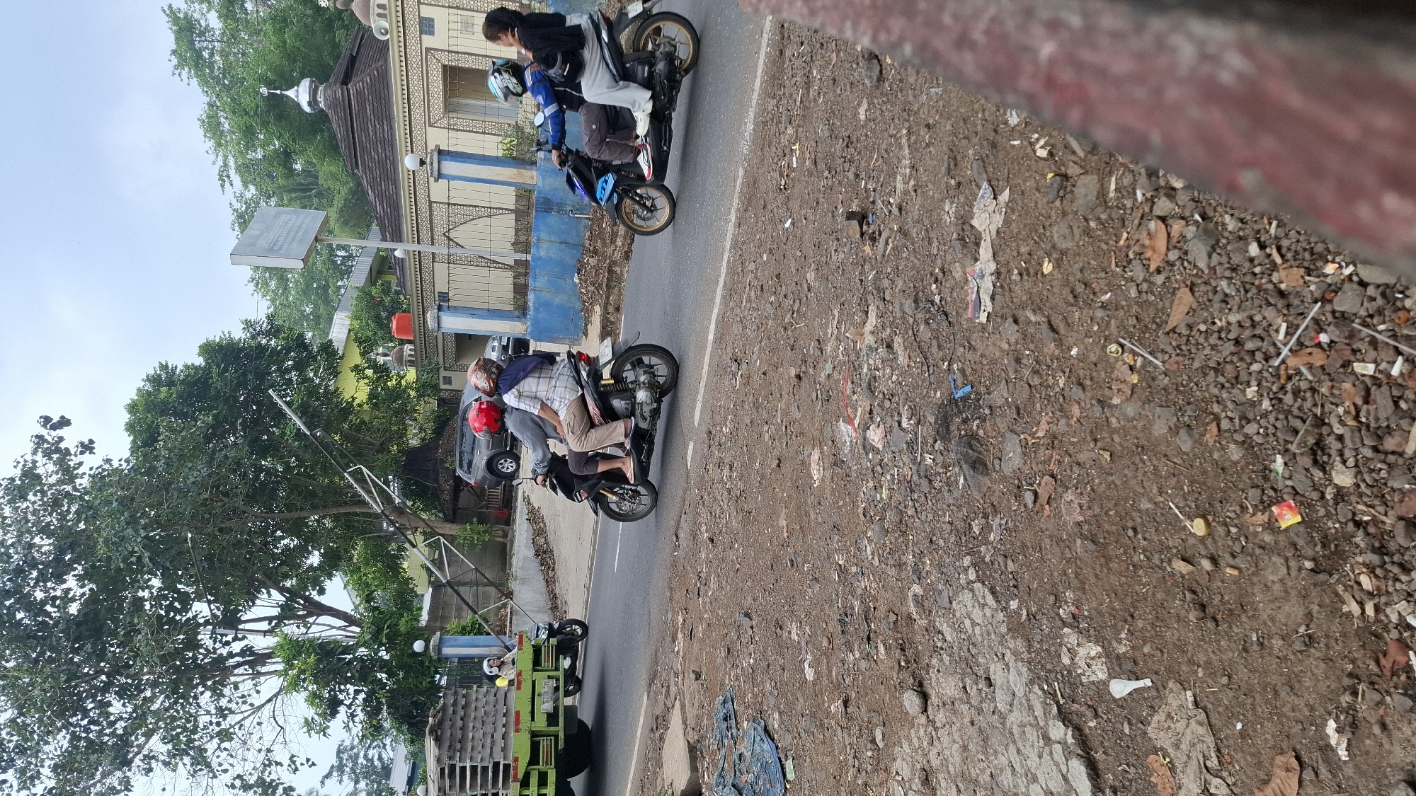 In the city of Cilaku, Indonesia, a scene unfolds on a paved road. The foreground is dominated by a rough, unpaved shoulder littered with debris, including small rocks, broken pavement, and various pieces of trash such as plastic wrappers and straws.
On the road, a green truck with a flatbed loaded with wooden pallets is parked on the left side of the frame, facing right. A person wearing a yellow shirt is seated on the truck. To the right of the truck, a motorcycle carrying three people is moving away from the viewer, also heading right. The driver wears a gray shirt and a red helmet. The passenger behind the driver wears a plaid shirt and a patterned helmet. The person at the very back wears a blue and white patterned headscarf and a plaid shirt.
Further to the right, another motorcycle travels in the same direction, carrying two people. The rider wears a blue jacket and a blue helmet, while the passenger, a woman, is wearing a black jacket and gray pants. Both are looking towards the right.
In the background, a building with a light yellow facade and a dark roof, possibly a mosque with a white dome and minaret, is visible. A sign is mounted on a pole in front of this building. A dark SUV is parked in front of the yellow building. Trees with lush green foliage line the background, suggesting a somewhat rural or suburban setting. The sky is overcast, contributing to the diffused lighting of the scene.