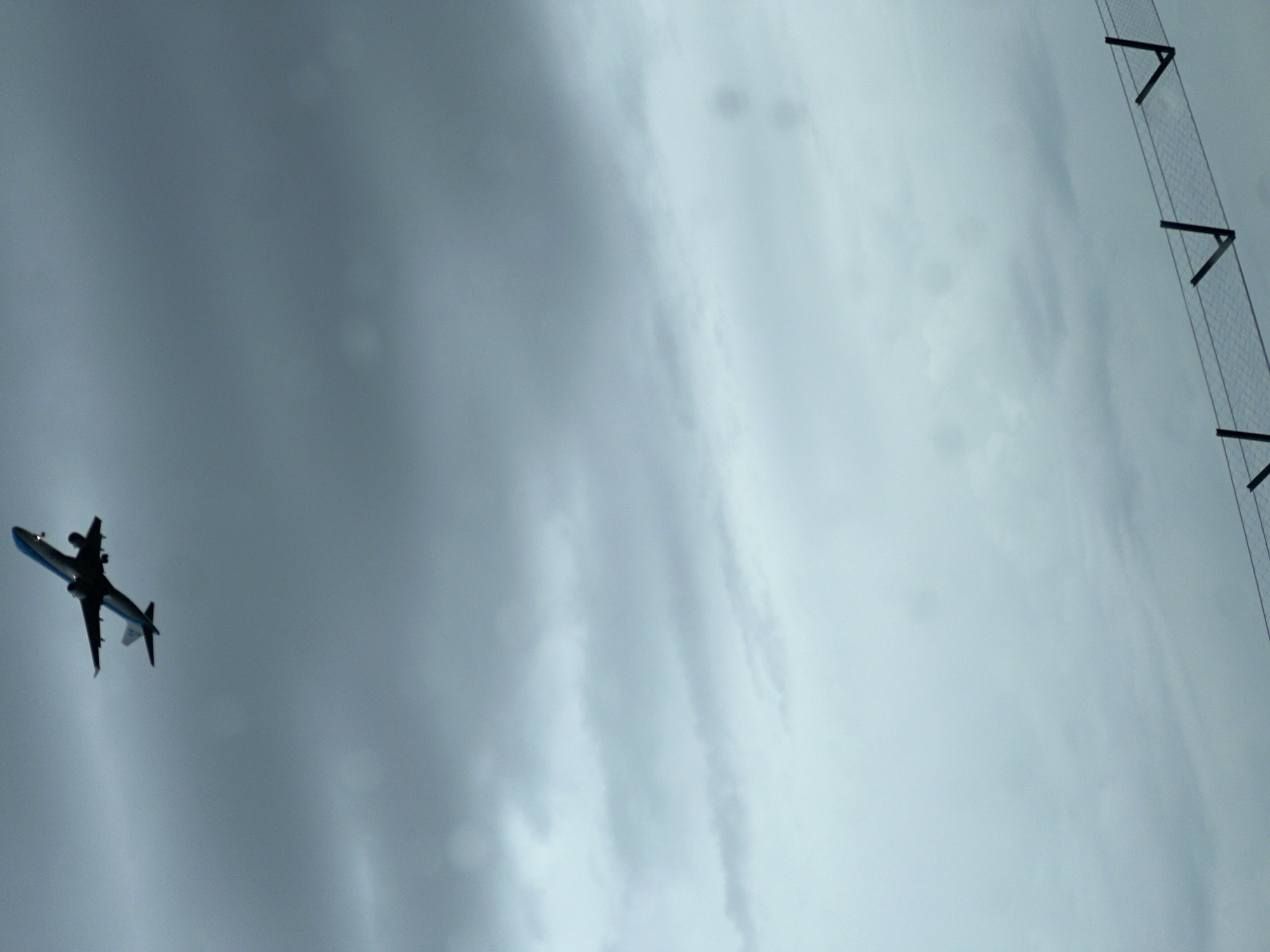 The image shows a low-angle view of an airplane in flight against a gray, overcast sky. A chain-link fence is visible in the upper right corner. It is impossible to determine the location of the image within Broadland, United Kingdom with the provided information. No people are visible.