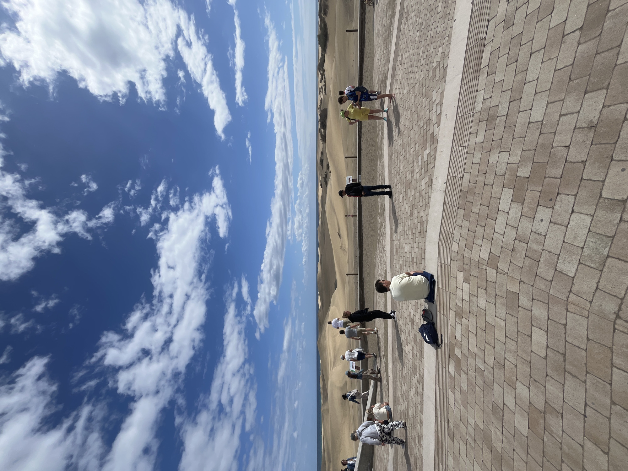 The image captures a scene in San Bartolomé de Tirajana, Spain. The view encompasses a blue sky with scattered white clouds, a sandy desert landscape, and a paved area. Several people are present, some standing and others walking along the paved walkway. One person is sitting. The paved area is constructed of rectangular stones. The desert landscape consists of rolling sand dunes.