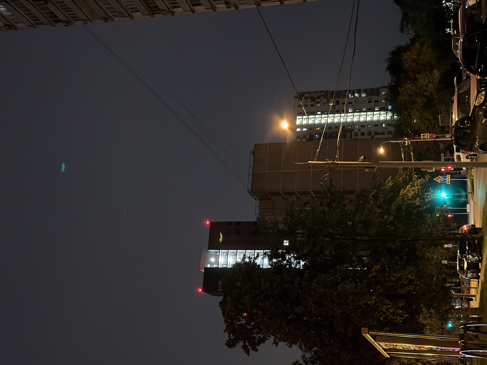 The image depicts a nighttime scene in a Moscow, Russia urban environment. The foreground features a street with several parked cars, illuminated by streetlights and traffic signals. Tall residential buildings with illuminated windows dominate the background, suggesting a densely populated area. Trees line the street, their foliage casting shadows in the dim light. Power lines crisscross the sky, a common feature of urban landscapes. The sky is dark and overcast, with a faint glow emanating from the city lights. A small, blurry light streak is visible in the upper portion of the sky, possibly from an aircraft or a natural phenomenon. The overall impression is one of a typical evening in a large city, with activity continuing after dark.