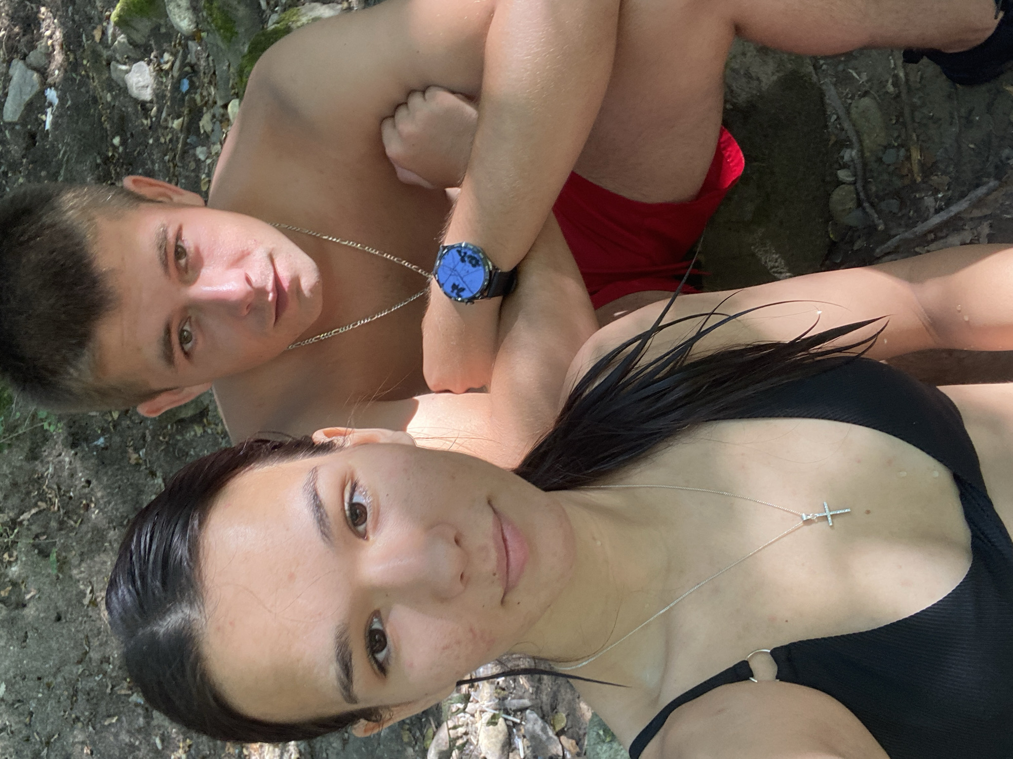 In a selfie taken outdoors, a young man and woman are positioned close together. The woman, in the foreground on the left, wears a black bikini top and a silver necklace with a cross pendant. Her dark hair is wet and pulled back into a ponytail, with strands falling to her shoulders. She has light skin and is looking directly at the camera. The man is to her right, shirtless, wearing red swim trunks and a silver chain necklace. He has short brown hair and is wearing a dark-faced watch on his left wrist. His arms are crossed in front of him, and he is also looking towards the camera. They appear to be sitting on the ground or a rock formation, surrounded by a natural environment with dirt, rocks, and foliage. The lighting suggests it is daytime, possibly a sunny day given the shadows and highlights on their skin. The setting suggests they are in a natural, outdoor location, possibly a riverbank or wooded area. Debnevo, Bulgaria is mentioned as the location.