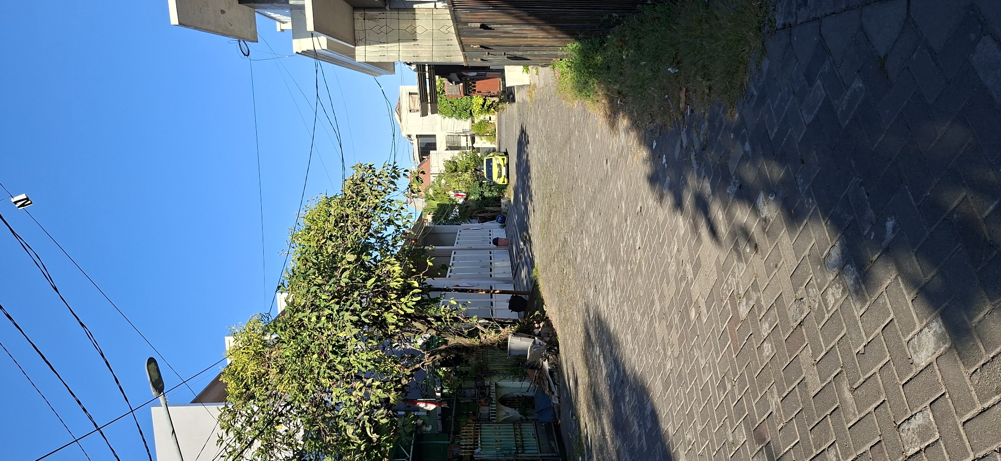 The image depicts a sunny street scene in Surabaya, Indonesia. The street is paved with dark gray interlocking bricks, and shadows from trees are cast across it. On the left side of the street, there is a large, leafy tree in front of a white fence and a modern-looking building. To the right of the tree, there are several trash bins. A green metal gate is visible further to the left, with some debris behind it.
On the right side of the street, there are more modern buildings with light-colored facades. A tall fence, possibly made of metal, runs along the edge of the property. Further down the street, a yellow car is driving away from the camera. The sky is clear and bright blue. Overhead, there are power lines and a street lamp attached to a pole on the left. A small white object with a black "N" on it is hanging from one of the power lines. The overall impression is a quiet residential street during the day.