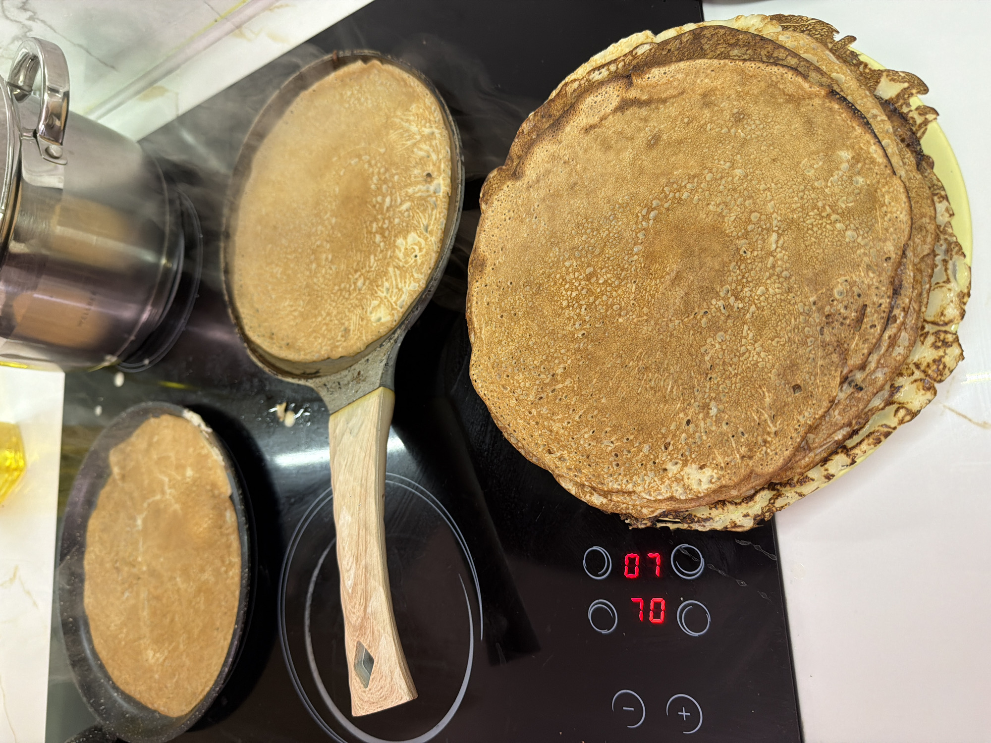 The image depicts a kitchen scene, likely indoors, focusing on the preparation of pancakes. There are three frying pans on a black stovetop. Two of the pans contain pancakes in the process of being cooked, with steam rising from one. The third pan is visible in the lower-left corner. A stack of cooked pancakes is piled on a plate to the right of the stovetop. A stainless steel pot sits on the left side of the stove. The stovetop's controls are visible, displaying the numbers "07" and "70." The location is not explicitly stated in the image but assumed to be Sochi, Russia.