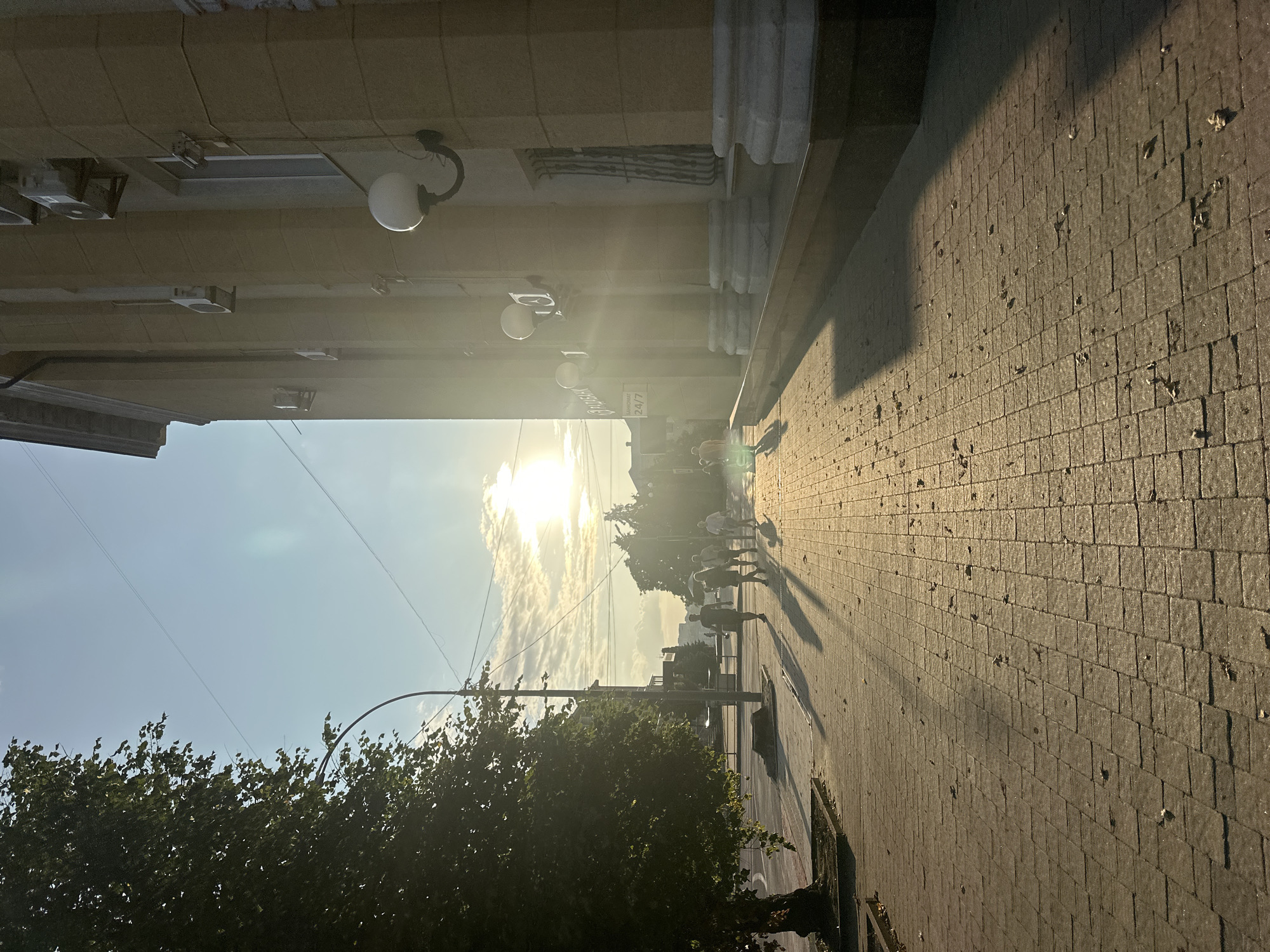 The image depicts a sunlit street scene in Ternopil, Ukraine, as indicated by the presence of a "24/7" bank sign written in Cyrillic script ("БАНКОМАТ 24/7"). The sun is low in the sky, casting long shadows across a paved sidewalk and street. A large tree with dark green foliage is prominent on the left side of the frame, with its shadow extending onto the sidewalk. A modern building with a light yellow facade, large columns, and air conditioning units is visible on the right. Several people are walking on the sidewalk in the mid-ground, casting distinct shadows. The sky is bright blue with some scattered clouds, and the sun's rays create a lens flare effect. The sidewalk is made of light-colored rectangular paving stones, with some fallen leaves scattered on it. A lamppost is positioned near the tree. The overall atmosphere is one of a warm, late afternoon in an urban environment.