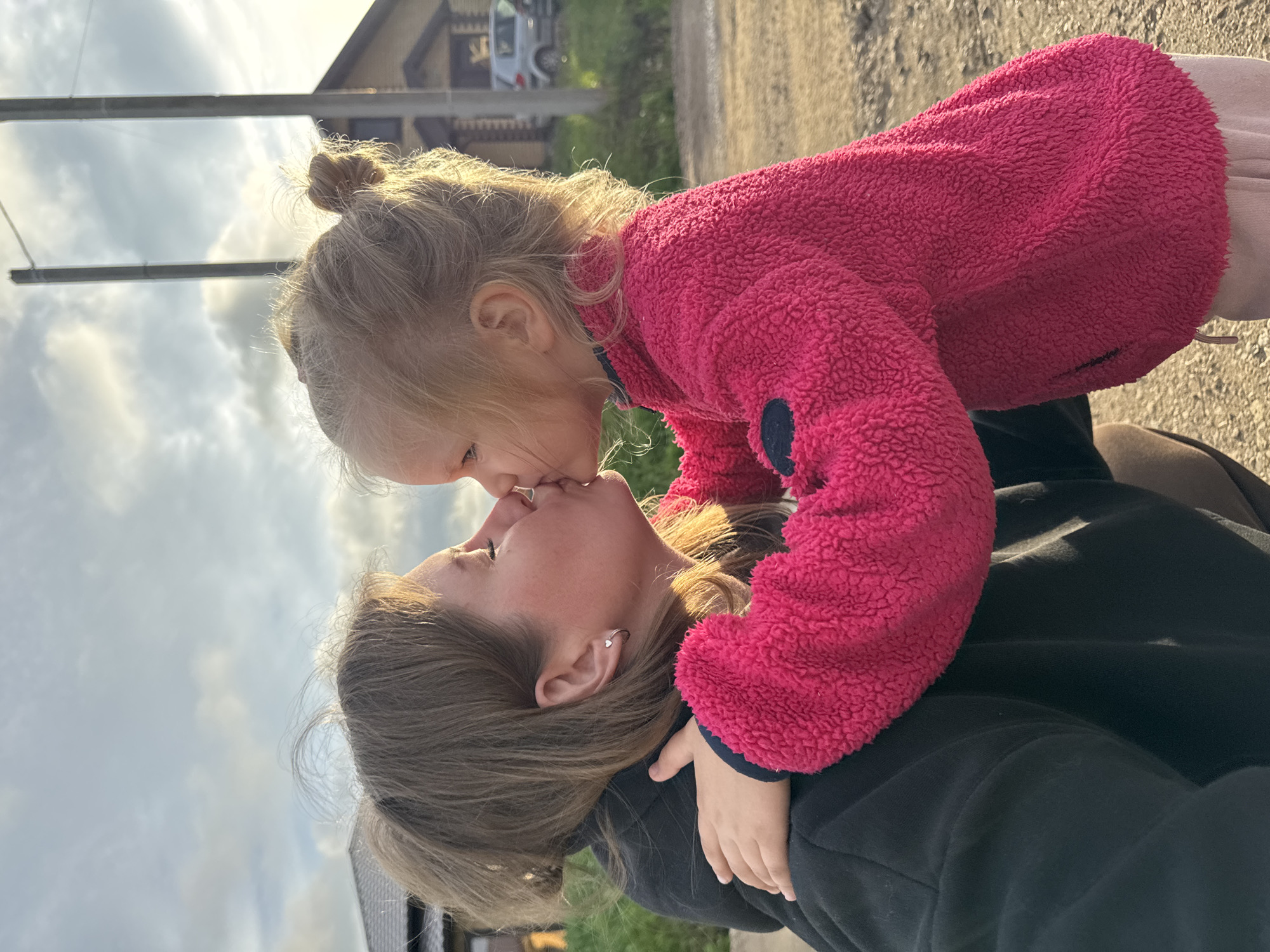 The image depicts an adult female and a young child engaged in a kiss, captured outdoors in Ферма, Russia, during late afternoon or early evening under a cloudy sky with prominent sunlight illuminating the subjects from the right.
The adult female, positioned on the left and leaning forward, presents with light brown hair, some strands visibly lifted against the sky. She wears a black hooded garment and a silver heart-shaped hoop earring in her left ear. Her eyes are closed during the interaction.
The child, a young girl estimated to be between two and four years old, is on the right, facing the adult. She has blonde hair styled in a small bun atop her head. She is dressed in a textured, fuchsia-colored fleece jacket with a dark circular patch on her left upper arm, and light-colored pants. Her left arm is wrapped around the adult female's neck or shoulder area.
The background reveals a semi-rural setting. To the left, a portion of a building with a dark roof and light-colored exterior walls is visible behind green vegetation. To the right, in the distance, a wooden house with dark trim is present, accompanied by two tall utility poles and a parked light-colored hatchback vehicle on an unpaved surface. The immediate foreground consists of dirt or gravel ground.