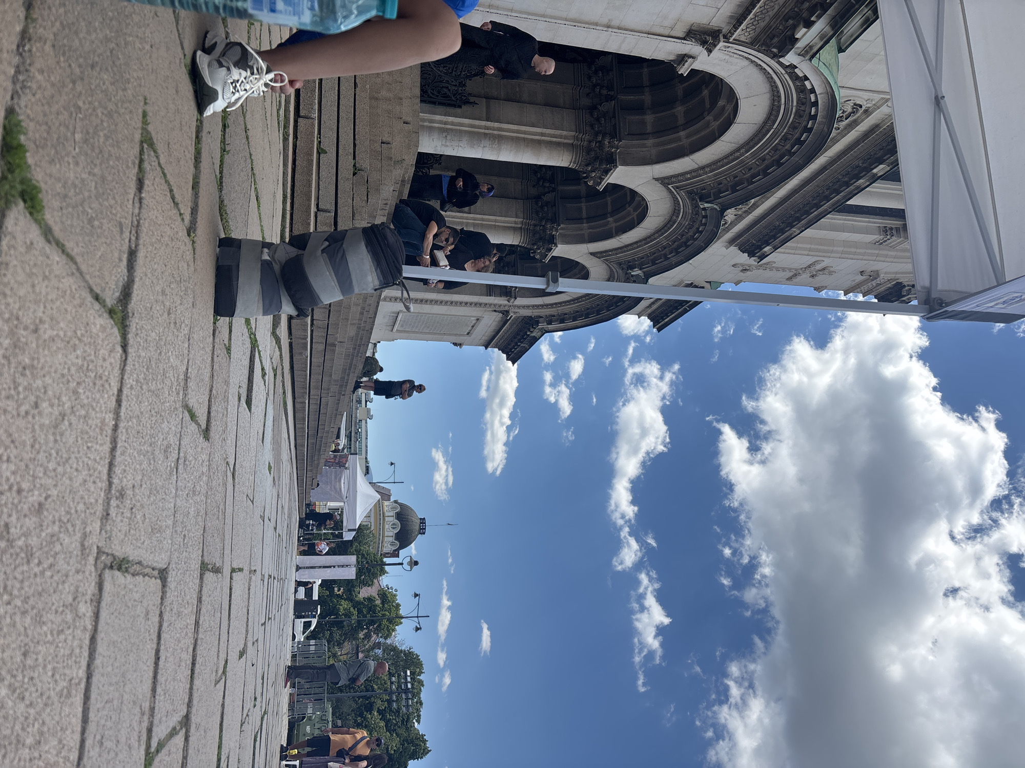 The image shows an outdoor scene in Sofia, Bulgaria, featuring the Alexander Nevsky Cathedral. Several people are present, some standing on the cathedral steps and others walking along the stone-paved walkway in front. A white tent-like structure partially obstructs the view on the right side. Above, a bright blue sky with fluffy white clouds dominates the background. Streetlights are visible, as are trees lining the walkway.