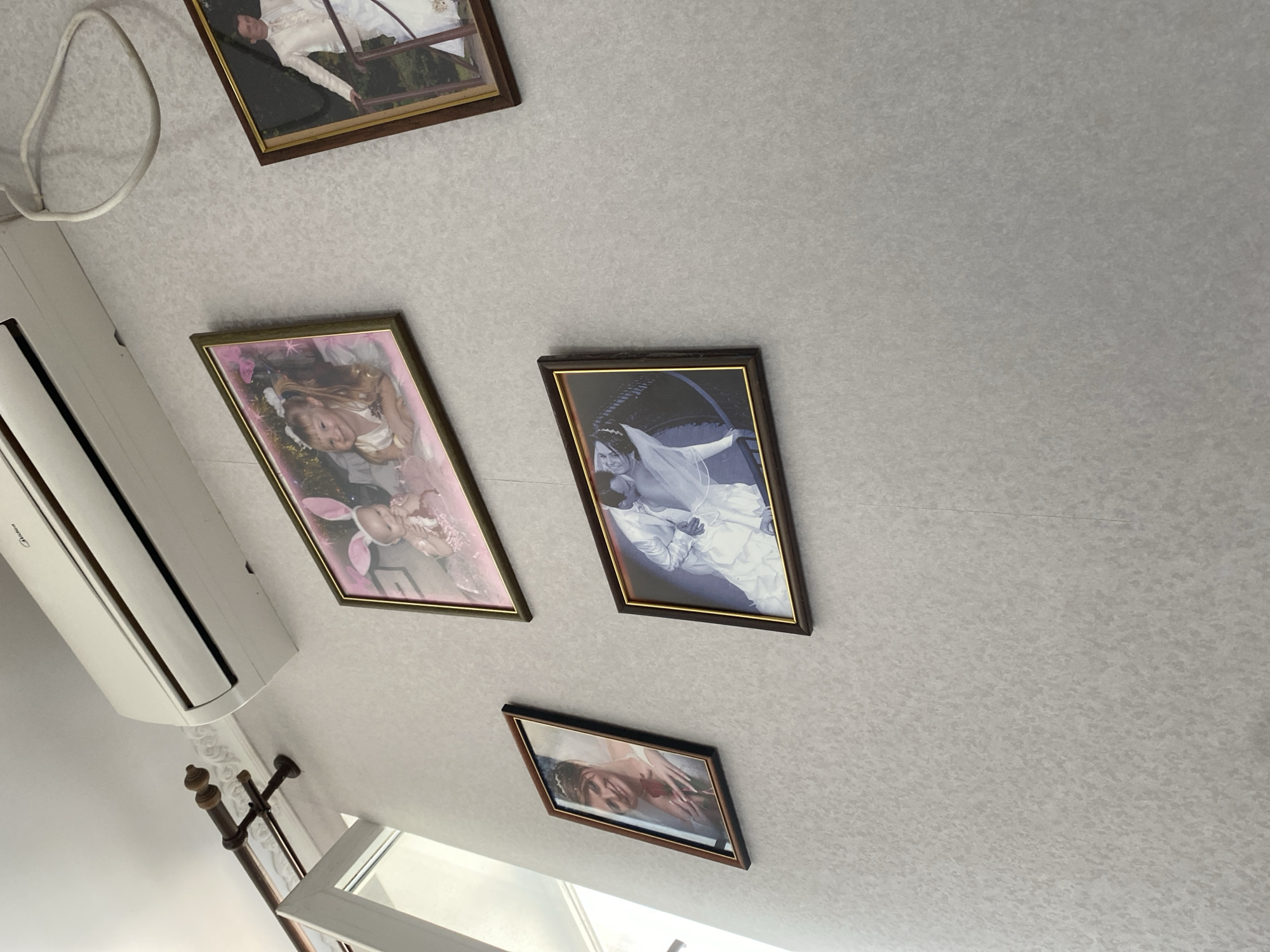 The image shows a section of a wall in an interior setting, likely a room in Molodizhne, Ukraine. The wall is covered in a light-colored, textured wallpaper. Mounted on the wall are four framed photographs.
The top left photo features an air conditioning unit mounted high on the wall, with a decorative curtain rod to its left.
The top middle photo is a portrait of two young girls, one wearing bunny ears and the other smiling.
The top right photo is a portrait of a man in a white suit.
The middle left photo is a portrait of a woman in a wedding dress holding a rose.
The bottom middle photo is a black and white image of a couple in wedding attire, with the groom kissing the bride.
The photographs are of varying sizes and orientations, hung at different heights on the wall, creating a gallery-like arrangement. The overall scene suggests a home or a communal space where family memories are displayed.