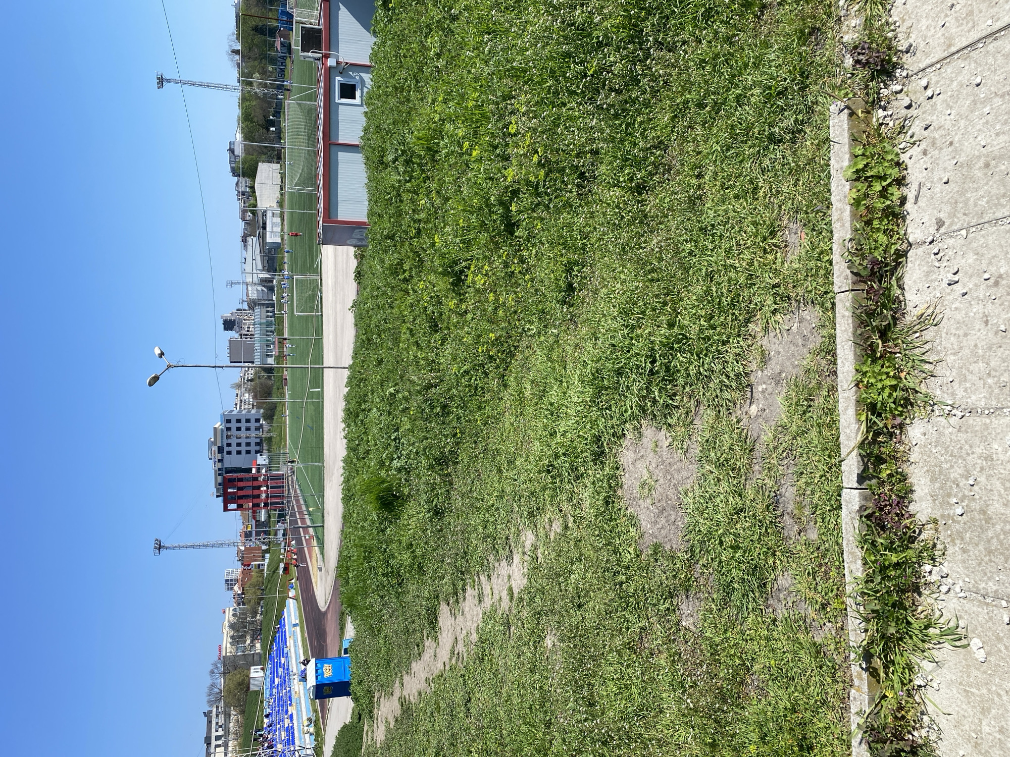 The image shows a view of the Spartak Stadium in Varna, Bulgaria. The stadium, with blue seating, is partially visible in the lower left portion of the image. Adjacent to the stadium is a running track (reddish-brown color). In the background are buildings and infrastructure, including what appears to be a sports field with goals and nets. The foreground primarily features a patch of grass and vegetation, running along the right side, bordered by a concrete walkway on the right edge of the image.