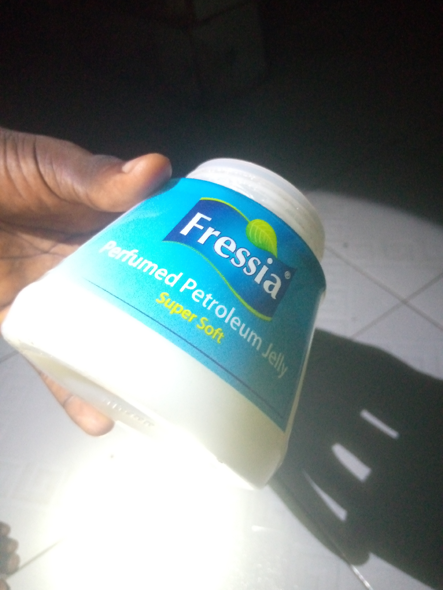 A hand holds a white plastic container of "Fressia Perfumed Petroleum Jelly Super Soft" against a dark background. The container has a light blue label with white text and a green leaf logo. The background appears to be a tiled surface with a bright light source casting a shadow of the container and hand.