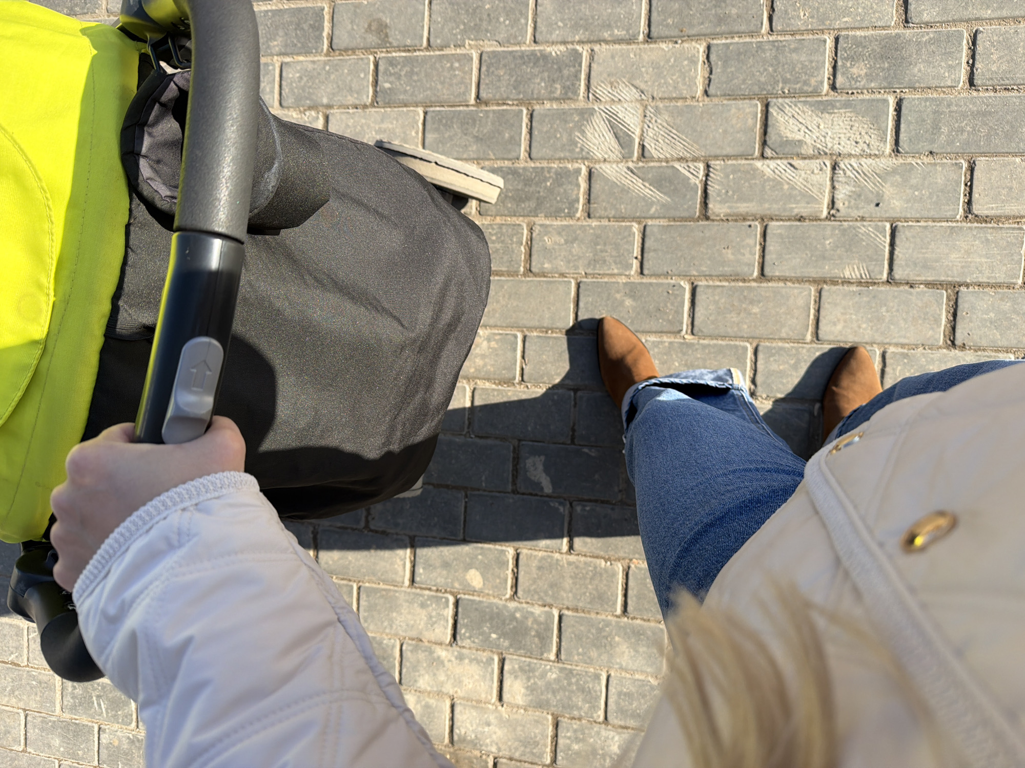 A high-angle shot shows a person walking with a baby stroller on a brick-paved walkway. The stroller is partially visible, featuring a bright yellow side and a black seat, with the person's hand holding the stroller handle. The person is wearing blue jeans and brown shoes, and a white quilted jacket. The ground is paved with gray bricks. The location is consistent with a pedestrian area in Votkinsk, Russia.