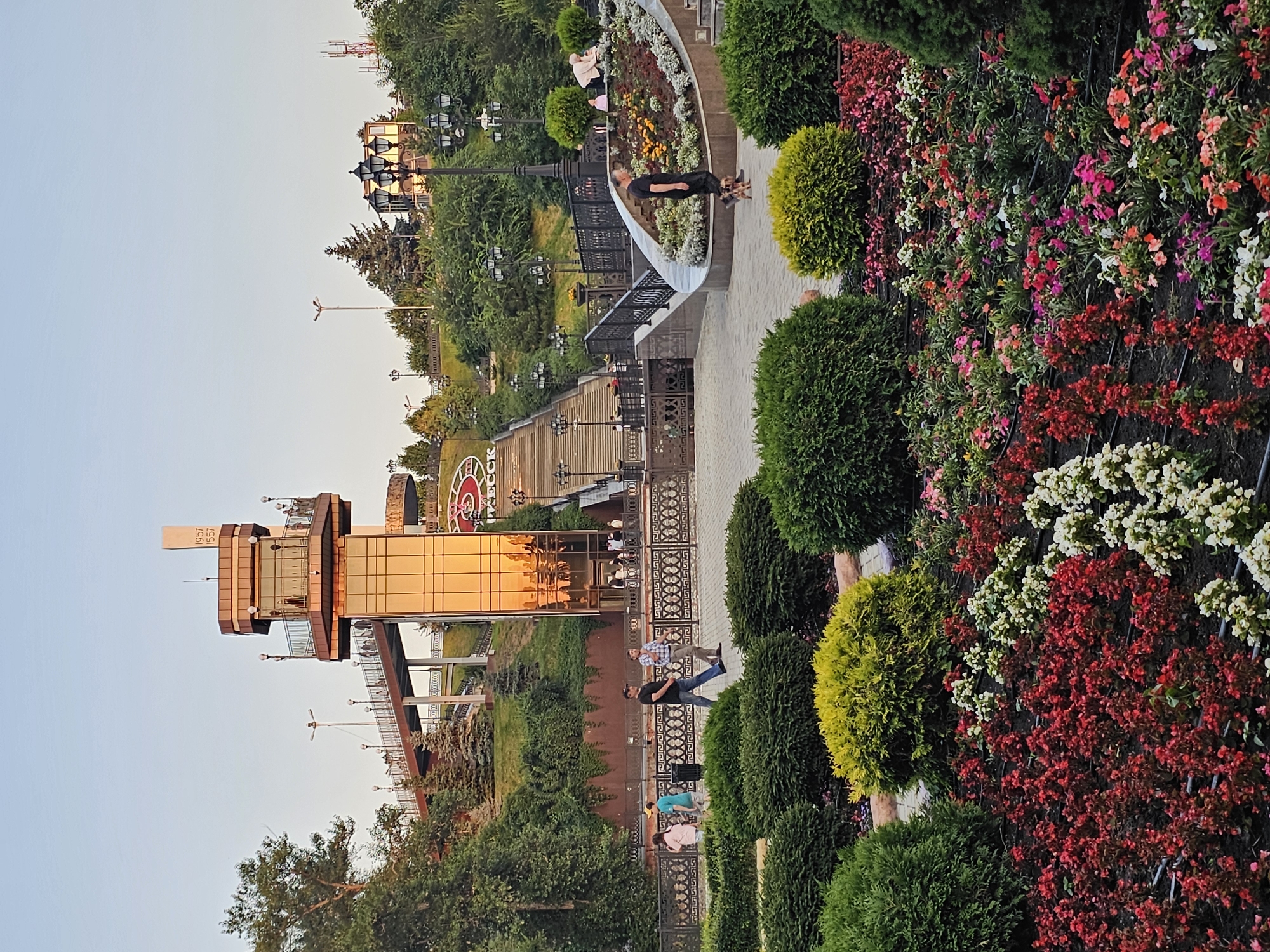 The image depicts a view of the city park in Cherkessk, Russia, featuring a prominent glass elevator tower and a grand staircase. The tower, with its reflective golden glass, stands tall against a clear sky. A walkway extends from the tower, leading to a bridge. The grand staircase, flanked by ornate lampposts and greenery, ascends the hillside. In the foreground, a vibrant flower bed is in full bloom with a variety of colorful flowers. Several people are visible in the scene: some walking on the paths, others on the bridge, and a man is seen walking his two small dogs. The overall scene conveys a sense of public space enjoyed during daylight.