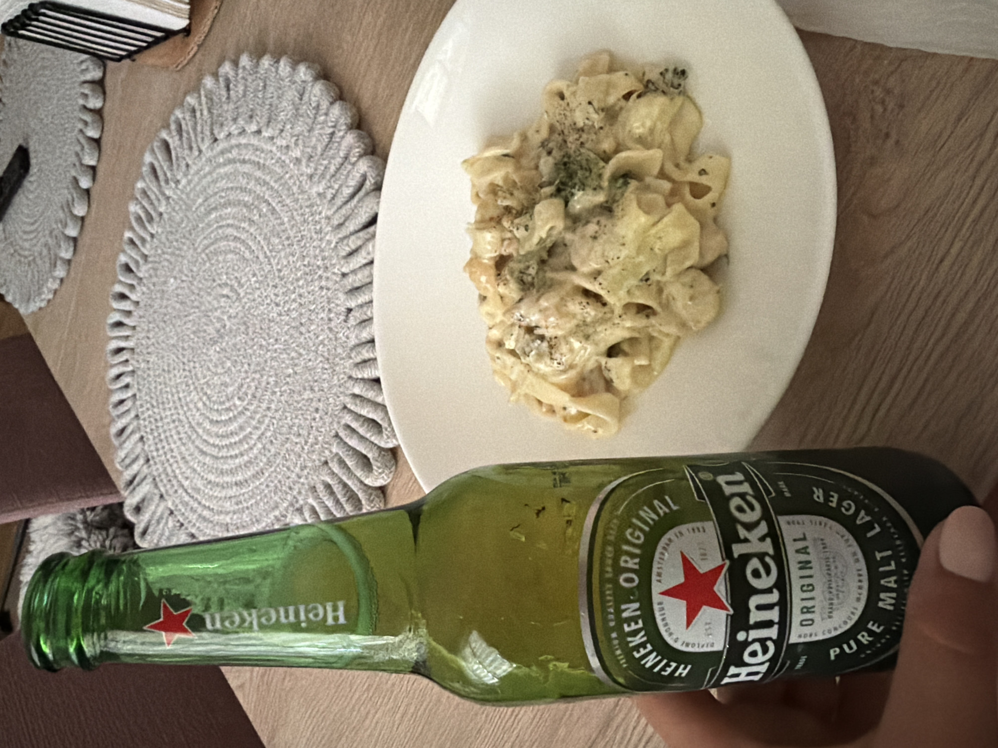 An overhead shot shows a hand holding a green Heineken bottle near a white plate of pasta. The pasta appears to be fettuccine with a creamy sauce, possibly with shrimp and broccoli. The background shows a wooden table with a grey circular placemat. The scene is indoors, likely a dining setting. The location is not specified in the image.