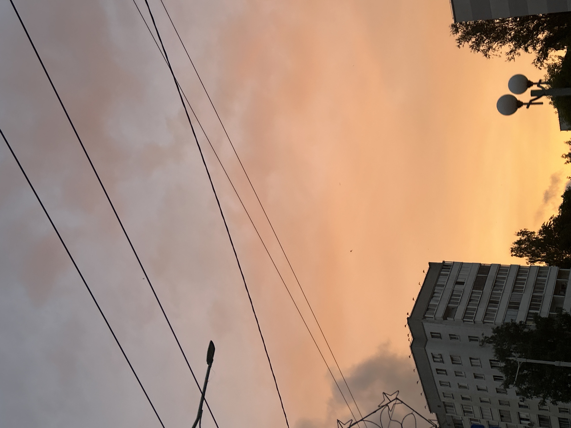 The image captures an urban scene during sunset in Minsk, Belarus. The sky is a gradient of pale purple to bright orange, with scattered clouds. Several black electrical wires crisscross the sky. In the lower left portion of the frame, the upper floors of a multi-story apartment building are visible. The building is grey and white with numerous windows and balconies. A street lamp is situated to the left of the building. To the right of the apartment building, the tops of two trees and a section of another building are visible. In the lower right quadrant, a street lamp with two spherical lights stands out against the sky. The overall impression is a quiet evening in a residential area of the city.