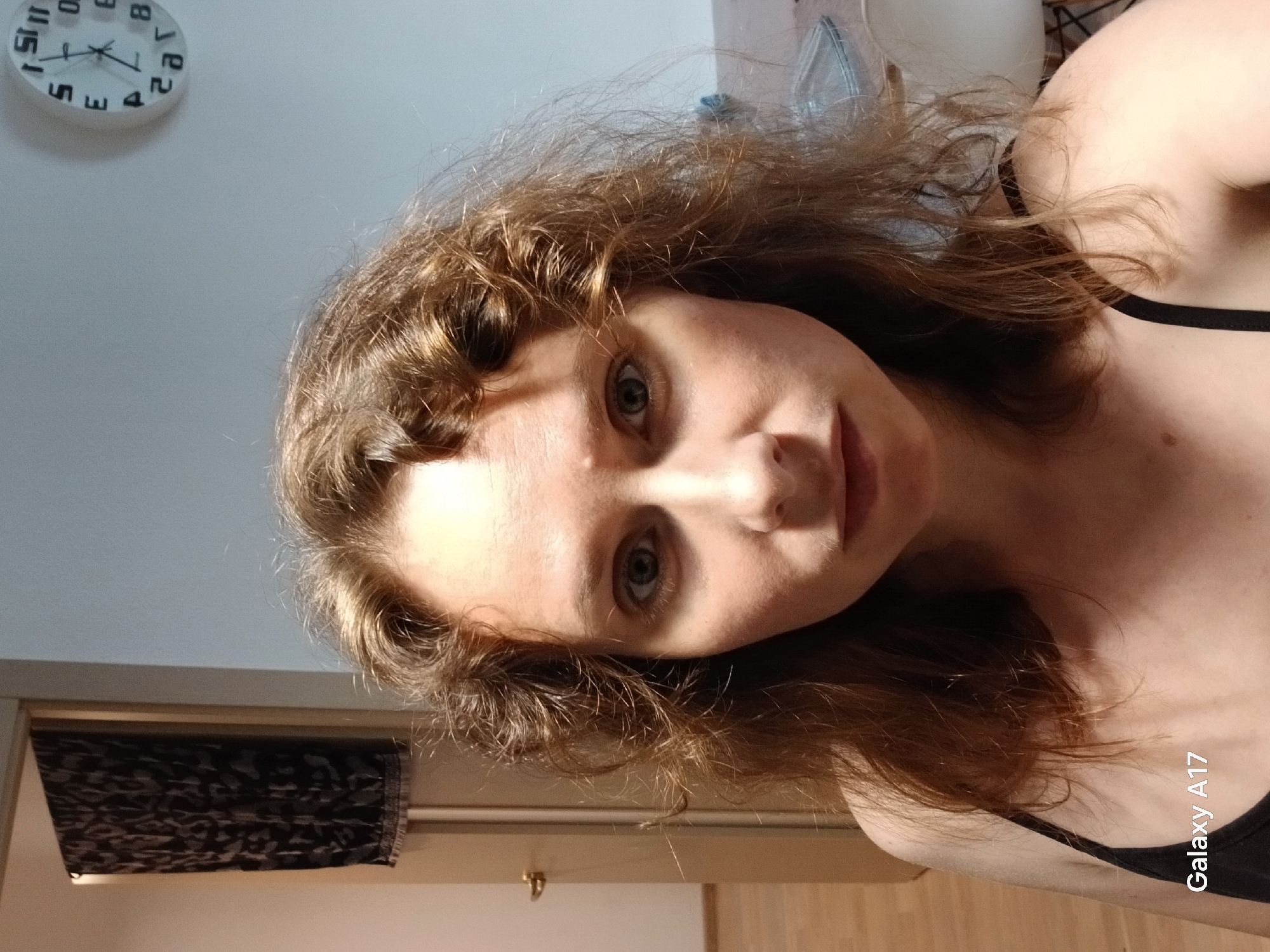 This portrait-oriented image, rotated 90 degrees counter-clockwise, depicts a young adult female from Yekaterinburg, Russia, looking directly at the camera. She has fair skin, light-colored eyes, and light brown, wavy, and somewhat disheveled hair that frames her face. A black strap, likely from a top or bra, is visible on her right shoulder/collarbone area. Her expression is neutral.
The background suggests an indoor setting, likely a home. In the upper left corner, a white circular wall clock with black numbers and hands is visible. The numbers appear mirrored, showing approximately 11:30 or 11:31. Below the clock is a light-colored wall. In the lower left, a dark, patterned fabric item, possibly a curtain or blind, is seen hanging. A wooden surface, which could be flooring or furniture, occupies the bottom portion of the image. The lighting appears soft and natural, illuminating the subject's face with some shadows on her right side. The photo seems to be a selfie due to the direct gaze and unusual, slightly upward angle. The text "Galaxy A17" is visible in white on the lower right side, indicating the device used to capture the image.