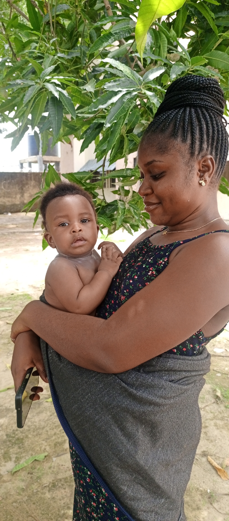 The image depicts a woman holding a baby in an outdoor setting. The woman has dark hair styled in braids, and she is wearing a floral patterned top. A gray shawl is wrapped around her torso, and she is wearing a necklace and earrings. She gazes at the baby in her arms. The baby is dark-skinned with short hair and appears to be unclothed from the waist up. The background includes a tree with green foliage, and a building is partially visible in the distance. The image was taken during daylight hours. Based on the prompt, the location is presumed to be Keffi, Nigeria.