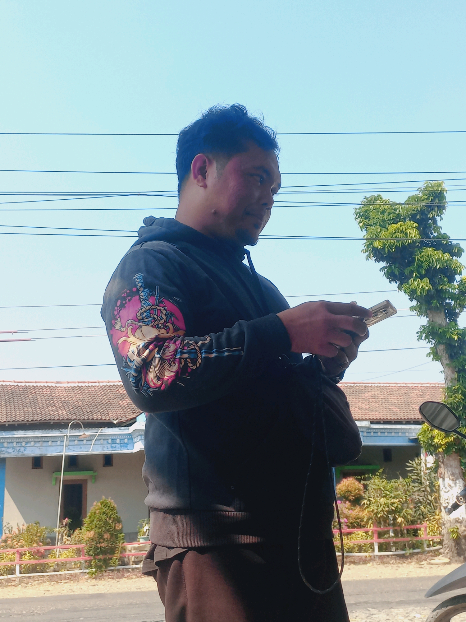 In Baturetno, Indonesia, a man stands outdoors against a clear blue sky, holding a smartphone in his hands. He is wearing a dark hoodie with a colorful, intricate graphic on the left sleeve, featuring what appears to be a dragon or mythical creature. The man's face is turned to his left, with a slight smile. His skin has a reddish hue on his cheeks and forehead, possibly from sun exposure or makeup. Behind him, a traditional Indonesian house with a tiled roof and blue trim is visible. The environment is arid, with dry ground and sparse vegetation. Power lines stretch across the upper portion of the image. To the right, a tree with lush green leaves stands tall. The scene suggests a casual moment captured in a residential area.