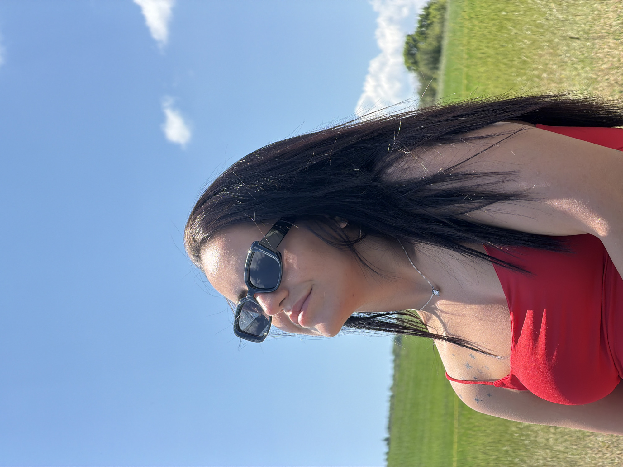 A young woman with dark hair and sunglasses, wearing a red top, poses in a grassy field under a clear blue sky with scattered clouds. The woman is looking upwards with her eyes closed, her face tilted towards the sun. A silver necklace with a small pendant is visible around her neck. She has small star-shaped tattoos on her left shoulder. The background consists of a vast green field stretching towards a treeline in the distance. The image is taken outdoors, likely in a rural area. The location is identified as Gurgulyat, Bulgaria.