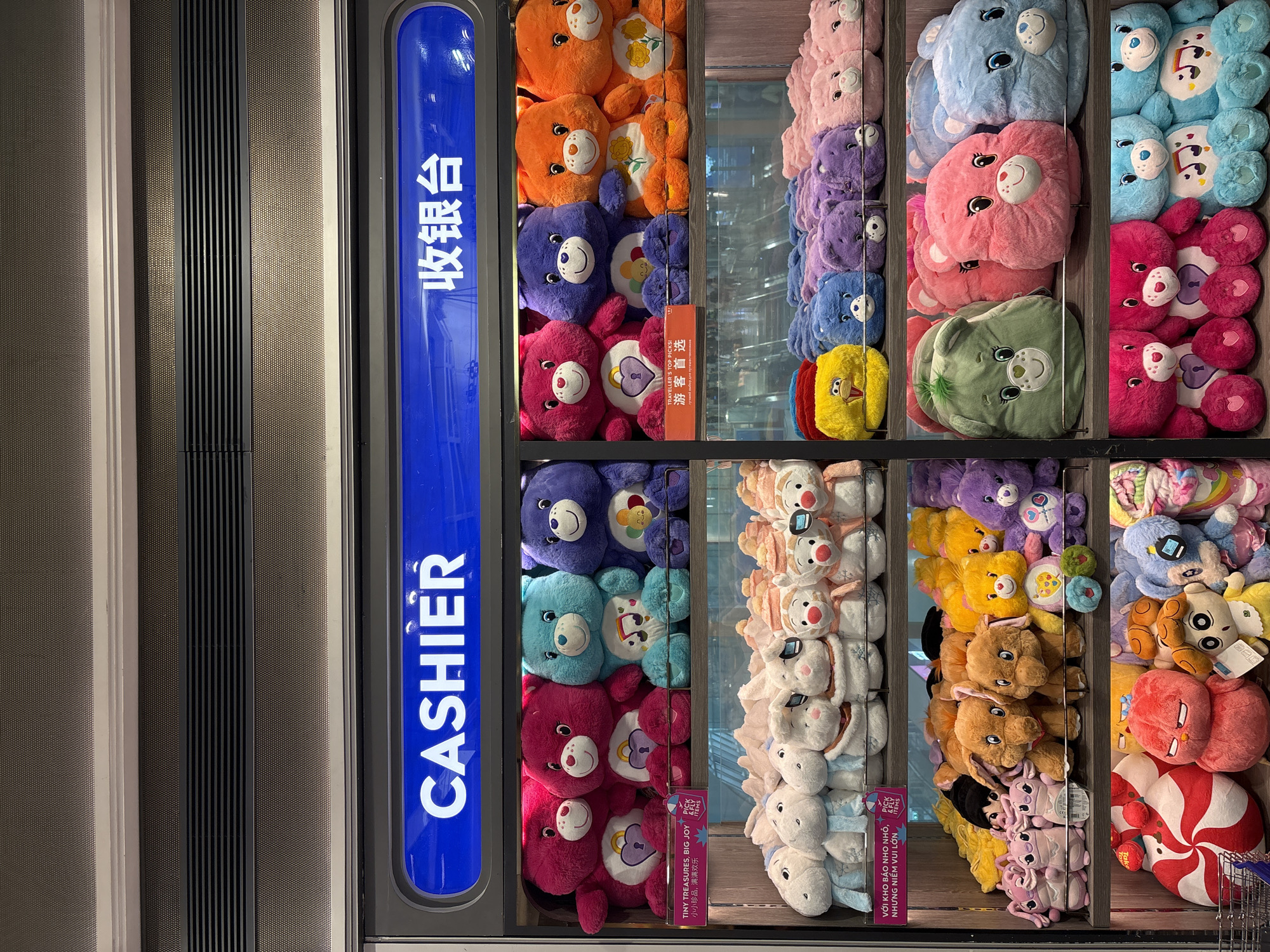 The image shows a display of plush toys in front of a cashier counter. The cashier counter has a blue illuminated sign with the word "CASHIER" in English on the left and Chinese characters on the right. The plush toys are arranged on shelves. The top shelf displays large, brightly colored plush bears, some in pink, teal, and purple. Below them, a shelf holds rows of smaller plush toys, resembling rabbits and other animals, in white and orange. The next shelf down contains a variety of plush toys, including more colorful bears in yellow, blue, purple, and pink, as well as some elephant-shaped plushies. The bottom shelf is filled with large, character-like plush toys, including a candy cane swirl, a pink bird-like creature, and other colorful figures. The overall environment appears to be an indoor retail space, possibly a gift shop or a store within an airport or train station.