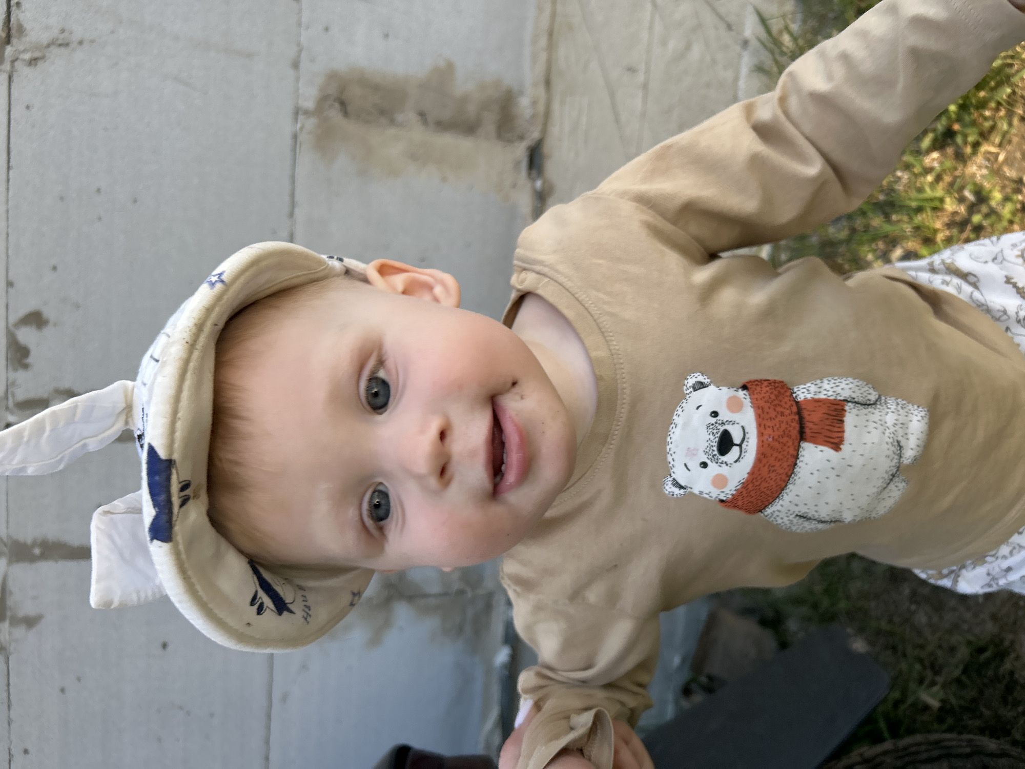 A young child, possibly a toddler, wearing a tan long-sleeved shirt with a polar bear design and a hat with floppy ears stands in front of what appears to be a concrete wall. The child is fair-skinned with light hair and blue eyes. The environment includes some greenery visible in the background, suggesting an outdoor setting. There is no information to confirm the location is in the city of Chaikovsky, Russia.