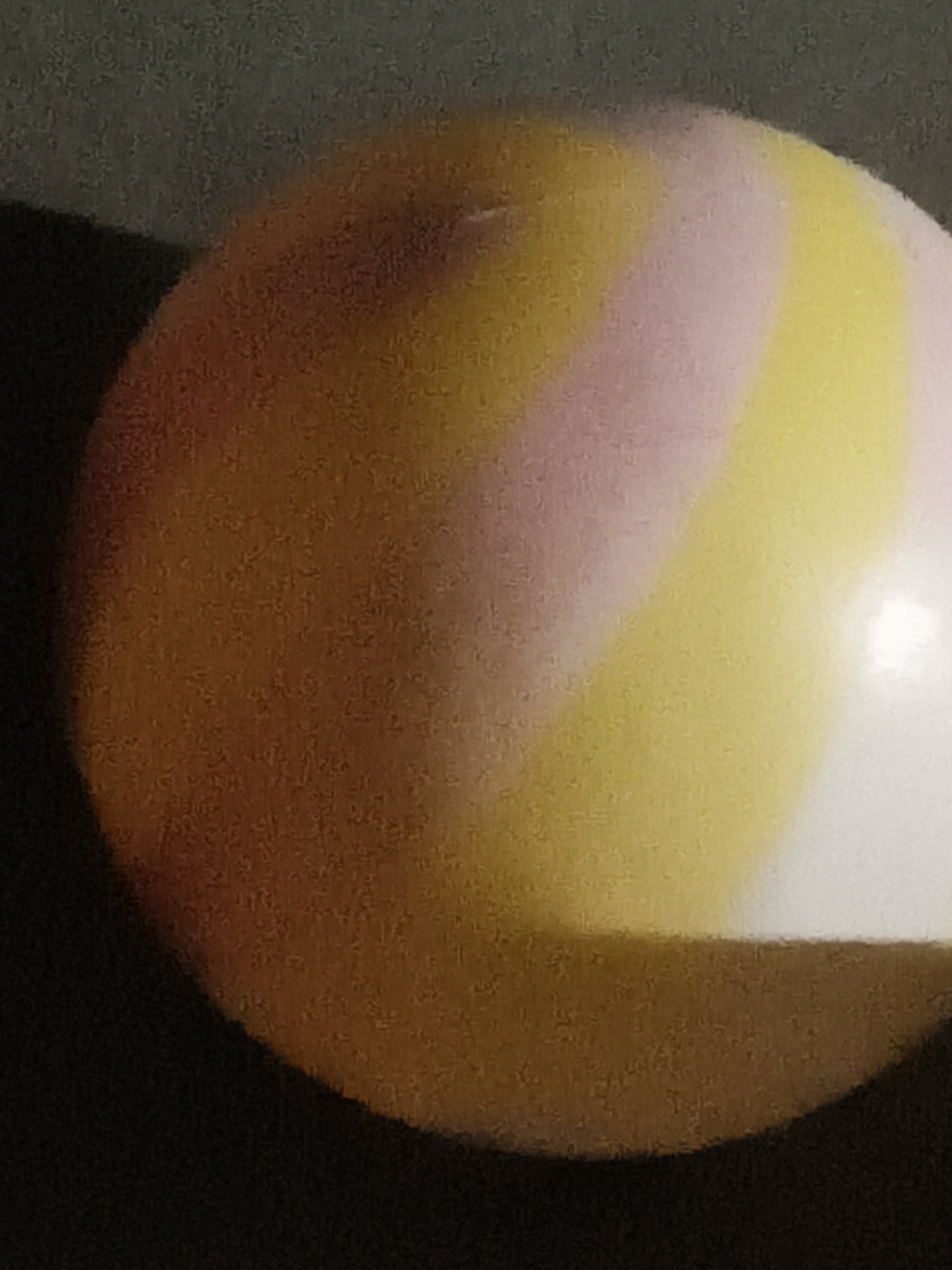 The image is a close-up shot of a ball with swirling colors of orange, yellow, and pink. The ball is spherical and appears to be smooth. The background is dark and out of focus, suggesting the ball is the main subject. There is a bright highlight on the right side of the ball, indicating a light source. The image quality is grainy. No specific location in Ubitpayaitek, Indonesia, can be determined from the provided image.