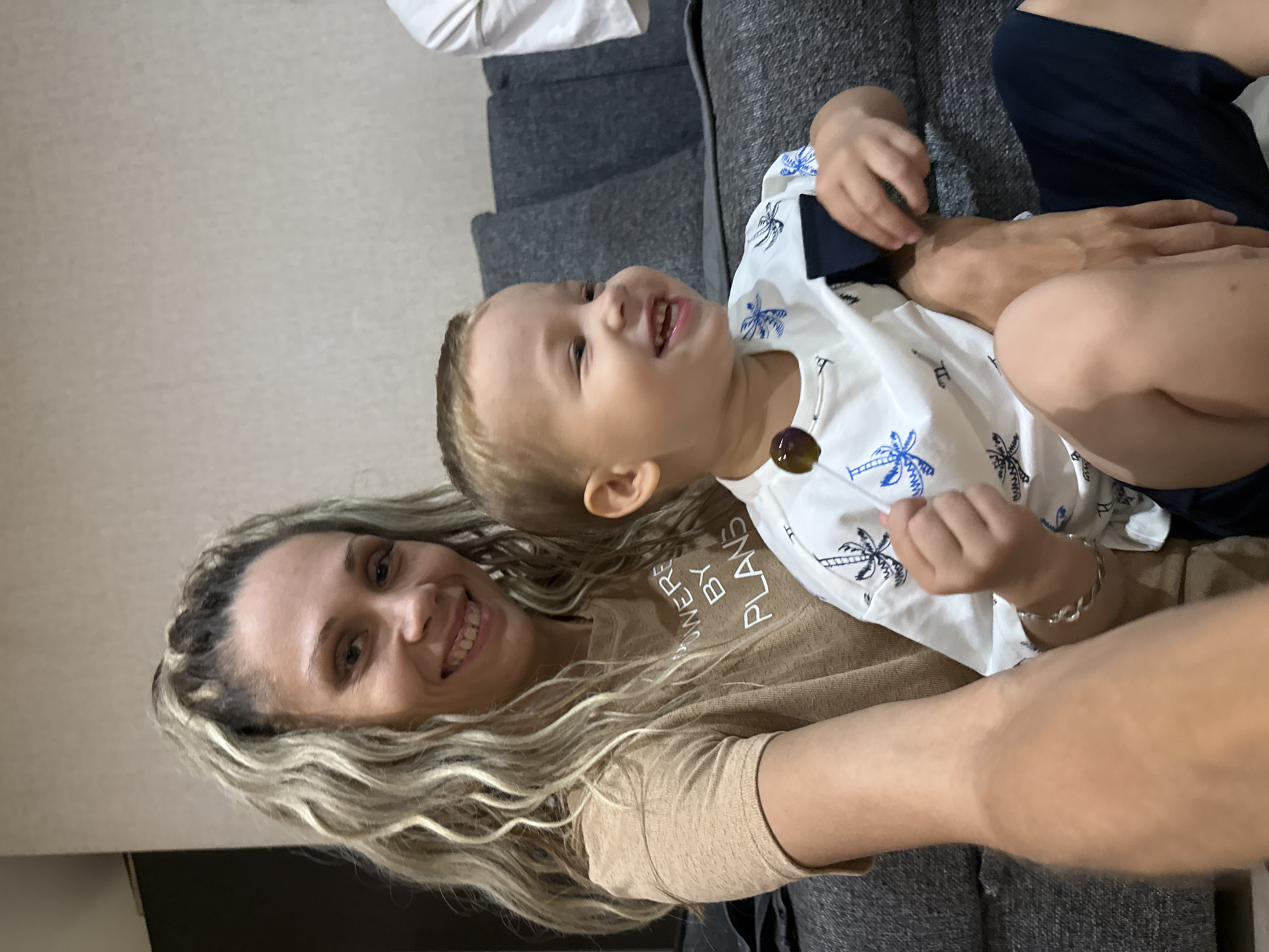 In an indoor setting, likely a living room, a woman with blonde, wavy hair pulled into a ponytail is seen from the chest up, holding a young child on her lap. The woman is wearing a tan t-shirt with white text that reads "WER E BY PLANS". She is smiling broadly, and her teeth are visible. The child, a boy with blonde hair, is facing to the right of the frame and laughing. He is wearing a white t-shirt with a blue palm tree pattern and appears to be holding a dark round object, possibly a piece of candy. The child's left arm is visible, and he is wearing a silver bracelet. The background consists of a dark gray couch with textured cushions and a light-colored wall. There is a hint of another person's white shirt visible on the far right of the image. The overall atmosphere is casual and familial. The location of Rusanovo, Russia, is not evident from the visual cues within the image.