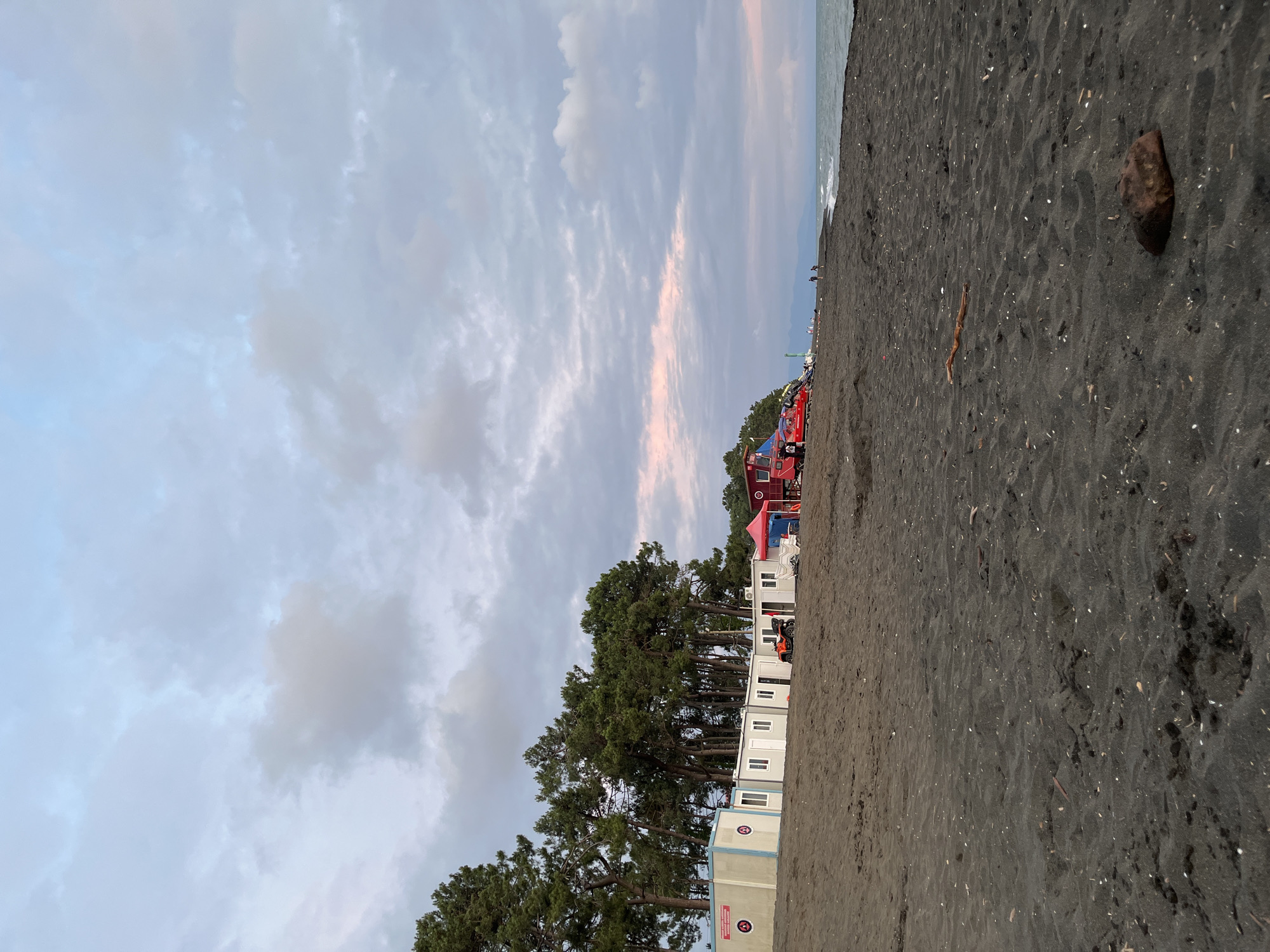 This is a low-angle, wide shot of a black sand beach in Ureki, Georgia. The sky is overcast with scattered clouds, tinged with pink and orange near the horizon, suggesting either dawn or dusk. A line of pine trees forms a backdrop to a row of modular buildings, some of which are white with blue trim. An orange ATV is parked in front of one of the buildings. Further down the beach, there are red structures, possibly lifeguard stations or rescue vehicles, and a small blue tent. A few distant figures can be seen walking along the shoreline. The sea is visible on the right, with gentle waves rolling onto the beach. The sand is dark and textured, with small shells and debris scattered across its surface. A rock is prominent in the foreground on the right.