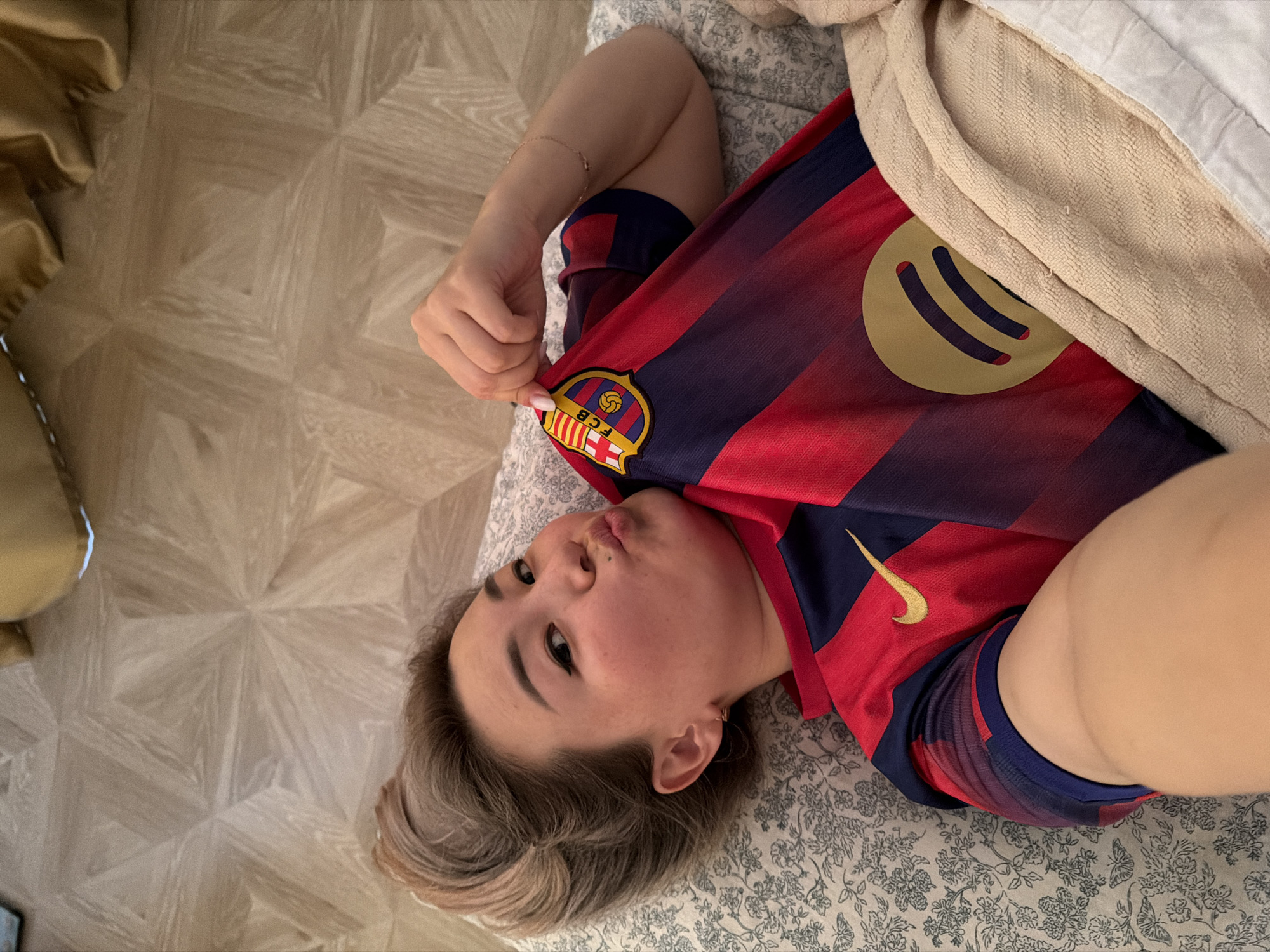 The image shows a person lying on a bed, wearing a red and blue striped jersey with a gold Spotify logo and the FCB crest. The person's head is turned towards the camera, and they are making a kissing face. The bed is covered with a floral patterned duvet and a beige blanket. The person's arm is resting on the jersey, and their hand is holding a corner of the shirt. The background shows a light-colored wooden floor with a geometric pattern. The lighting suggests it is daytime. The person has short, blonde-highlighted hair and is wearing subtle makeup, including eyeliner. A thin gold bracelet is visible on their wrist. The overall impression is casual and playful.