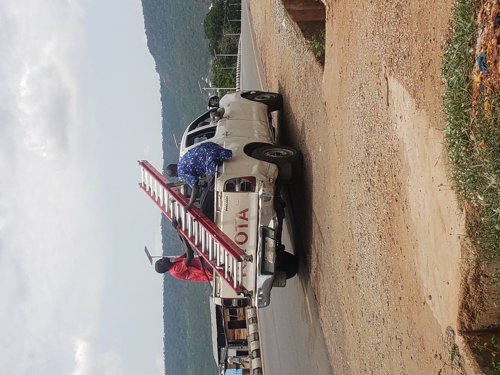 Here is a description of the image:
The image depicts a white Toyota Hilux pickup truck parked on the side of a road in Adankolo, Nigeria. Two men are on the truck with a red ladder. One man in a blue patterned shirt is leaning into the cab of the truck, while another in a red shirt is standing on the rear of the truck, holding the ladder. The environment includes a road with a yellow line, mountains in the background, and vegetation along the roadside.