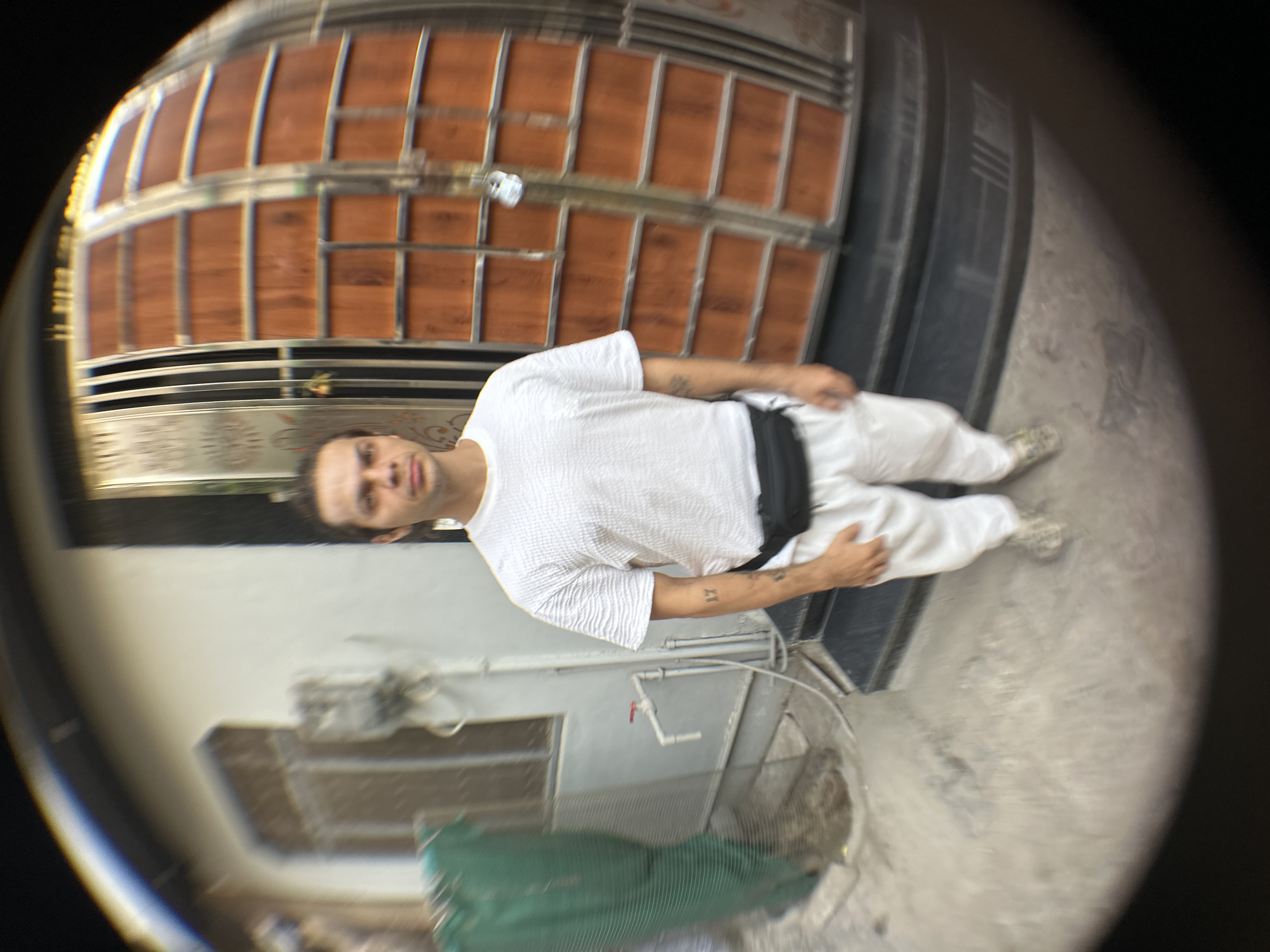A full body shot of a man in white clothing is captured through a fisheye lens. The man, with dark hair pulled back, stands in front of a brown and silver gate. He wears a white short-sleeved shirt and white pants, with a black fanny pack around his waist. Tattoos are visible on his left forearm. The background includes a light grey wall, pipes, and part of a green object. The location is identified as Mathura, India.