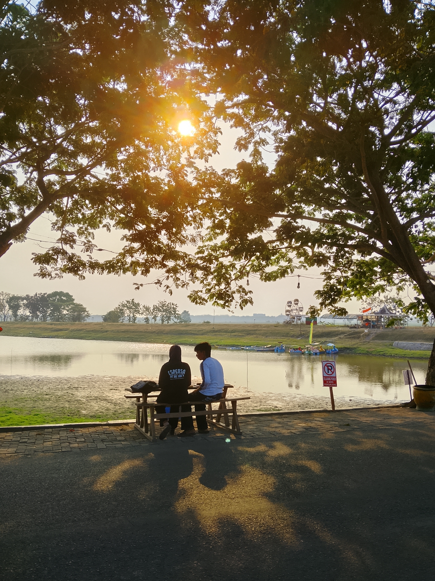 Two individuals are seated at a wooden picnic table near a body of water. The person on the left wears a black hooded sweatshirt with white text. The person on the right wears a white long-sleeved shirt. They are positioned facing away from the camera, looking towards the water. The sun is shining brightly, casting warm light and dappled shadows across the scene. The water is calm, reflecting the sky. In the background, across the water, there is a Ferris wheel, several small boats, and structures that appear to be part of an amusement or recreational area. The environment is outdoors, under the canopy of large trees. The ground in front of the table is paved with interlocking stones. A red and white sign with a circled "R" and text in Indonesian is visible on a post near the water's edge. The overall scene suggests a peaceful outdoor setting, possibly a park or recreational lake, in or near Ngadrin, Indonesia.