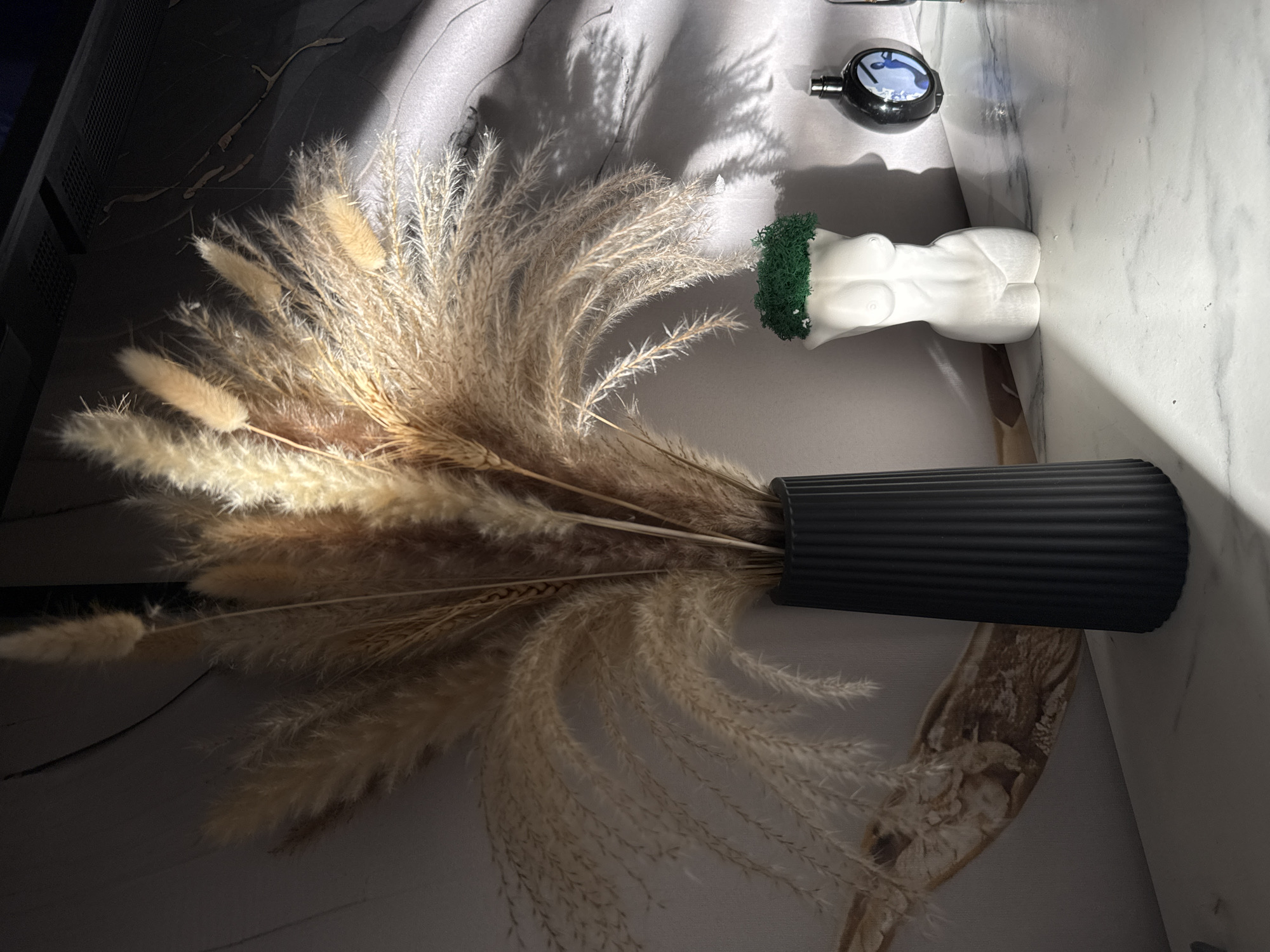 The image displays a still life arrangement on what appears to be a marble or marble-patterned surface. A black, ribbed vase holds a large bunch of dried pampas grass, which spreads out across the surface. Adjacent to the vase is a small, white anatomical sculpture of a female torso with green moss or similar material around the neck area. A small, black and silver object, possibly a decorative item, sits near the sculpture. The lighting is uneven, with strong shadows and highlights, suggesting a single light source, possibly sunlight. The photograph was taken indoors. There is no indication this photo was taken in the city of Chernushka, Russia.
