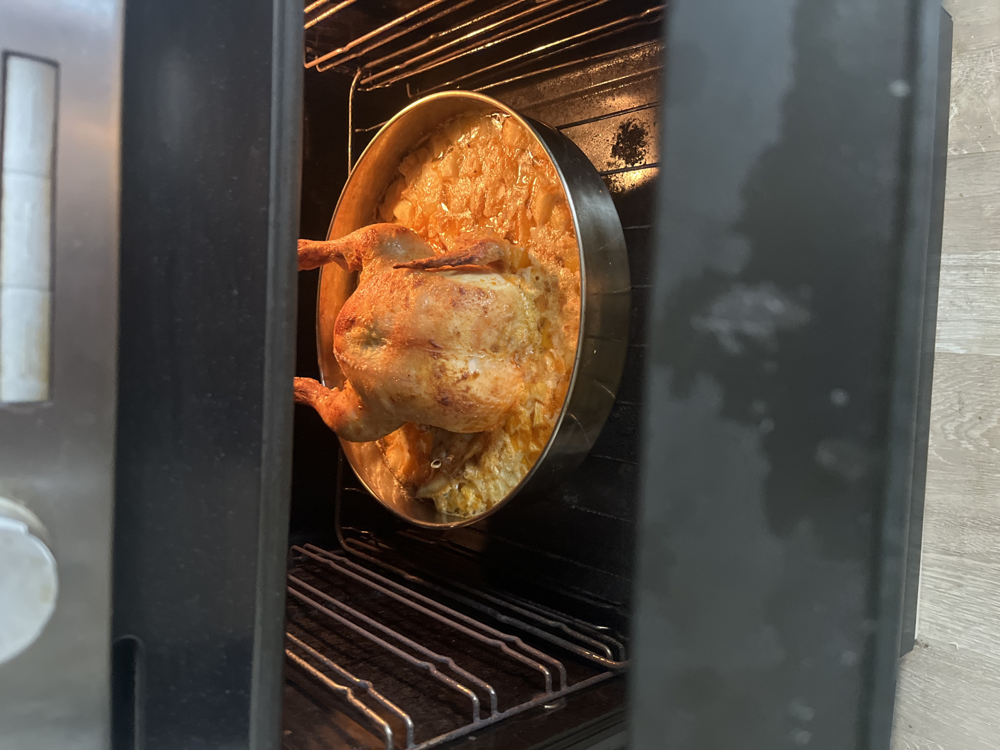 A cooked whole chicken with potatoes is in a round, silver pan inside an oven. The oven door is ajar, revealing the oven rack below the pan. The oven appears to be located indoors. There is no indication of Burgas, Bulgaria.
