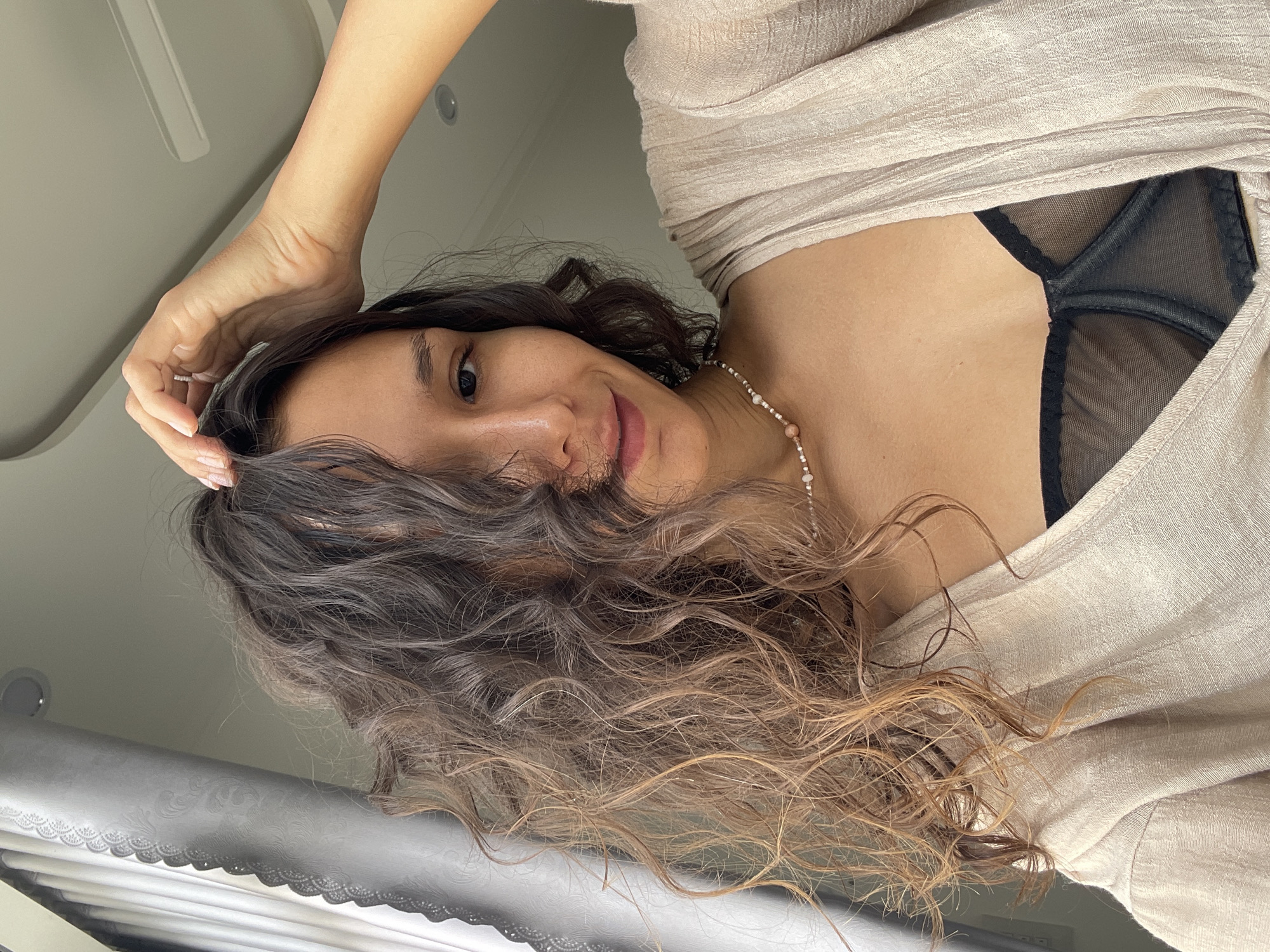 A person, likely female, is featured in a close-up, low-angle shot, looking directly into the camera with a gentle smile. They have long, wavy, dark hair that transitions to lighter brown or blonde at the ends, framing their face. One hand, adorned with a simple ring, rests on their head, fingers entwined in their hair. The subject is wearing a light beige, loose-fitting top, revealing a black, sheer-patterned bra underneath. A delicate necklace composed of small white and pinkish beads is visible around their neck.
The background suggests an indoor setting, with a light-colored ceiling visible at the top, featuring a circular light fixture or vent. Below the subject, a white surface with a decorative, scalloped pattern, possibly a blind or curtain, is partially visible. The lighting is soft and ambient, characteristic of an indoor environment. The activity depicted is posing for a photograph. No text is visible in the image. The provided context indicates the location is Mũi Né, Vietnam.
