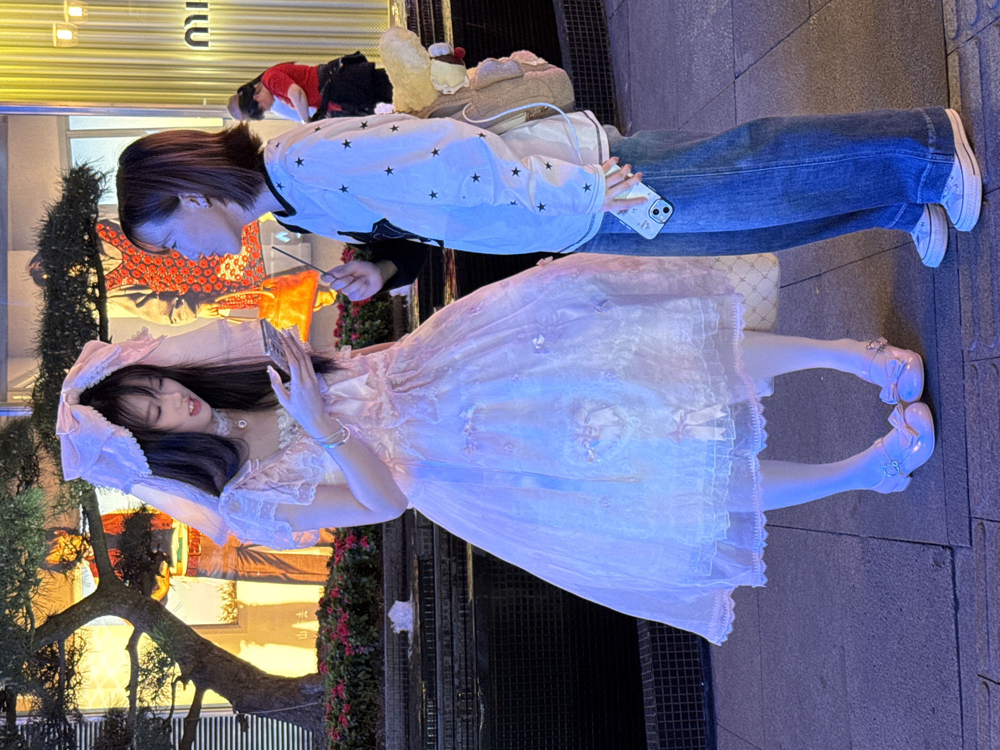 On a sidewalk in Yuzhong District, China, a young woman in a hooded light pink frilly dress and pink shoes, is posing while looking at her cell phone. A second person with short dark hair and wearing a white shirt with black stars, blue jeans, and white sneakers is using their cell phone to take a photo of the first woman. The scene also includes a low tree, a decorative bush, and an outdoor storefront.