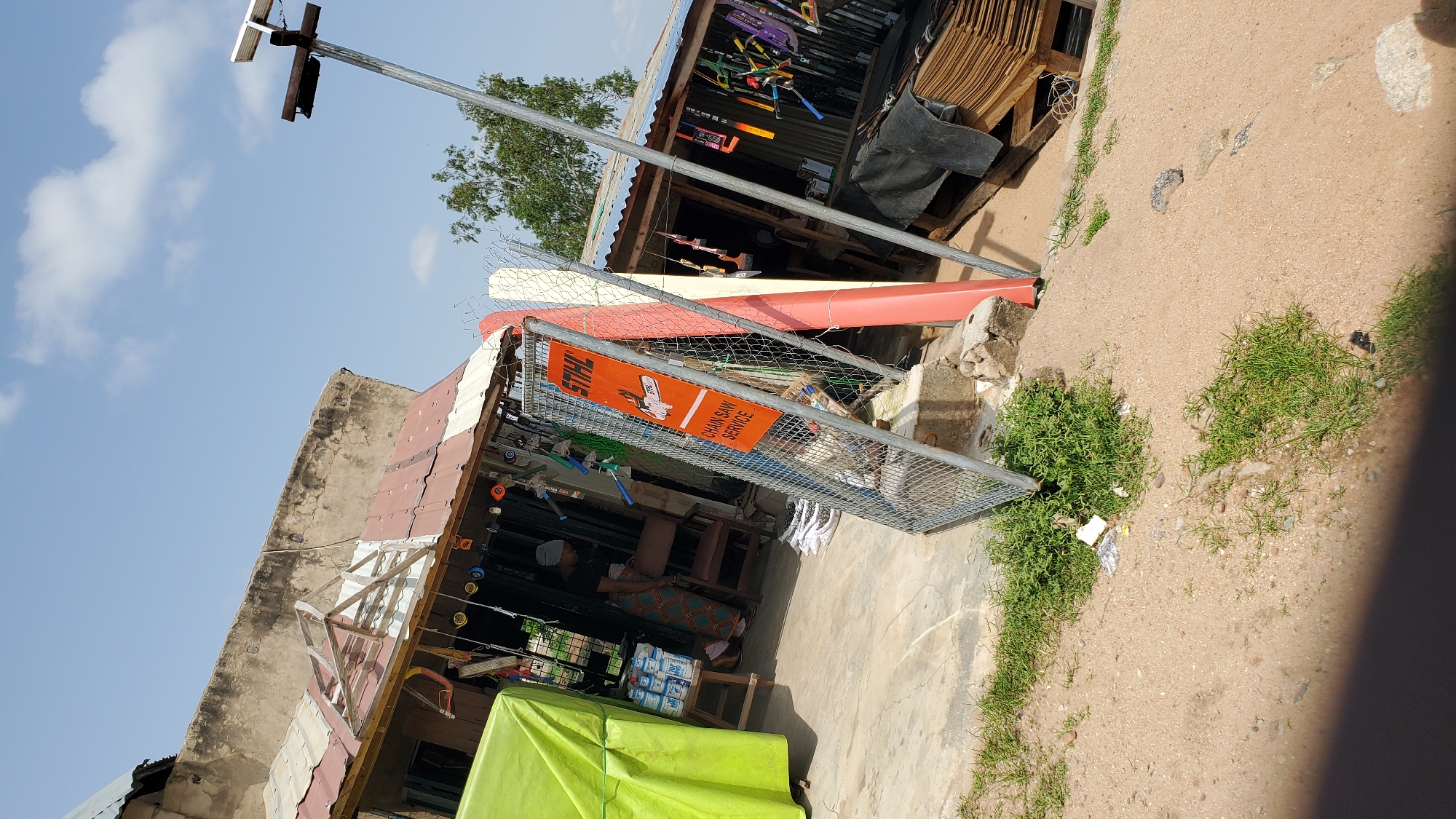 An outdoor scene depicts a small shop with a tin roof in Jalingo, Nigeria. The shopfront is cluttered with various items, including a bright green tarp on the left, several cans stacked on a wooden stool, and a woman in a patterned skirt standing inside. Hanging from the shop's awning are numerous colorful objects, possibly tools or accessories. In front of the shop, a metal fence encloses an orange sign that reads "STIHL CHAIN SAW SERVICE". To the right of the fence, two tall, thin objects, one red and one white, lean against the shop. The ground is a mix of sand and sparse green grass, with some scattered debris. A tall metal pole with a solar panel is visible in the background, along with trees and a clear blue sky with some clouds. The scene is illuminated by bright sunlight, casting shadows.
