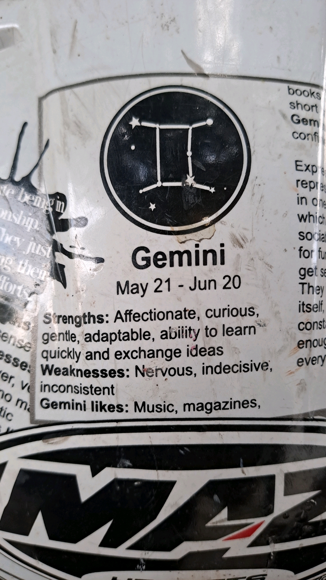 The image is a close-up of a sticker with astrological information for Gemini. The sticker features the Gemini constellation symbol within a black circle. Text below the symbol details Gemini's dates (May 21 - Jun 20), strengths (affectionate, curious, gentle, adaptable, ability to learn quickly and exchange ideas), and weaknesses (nervous, indecisive, inconsistent). It also lists Gemini's likes (music, magazines). The sticker is applied to a textured, possibly plastic surface, with visible scratches and smudges. The background is a light color, and part of a black and white logo is visible at the bottom of the frame. There are no people or specific locations in the city of Julicotmeurak, Indonesia, depicted in this image.