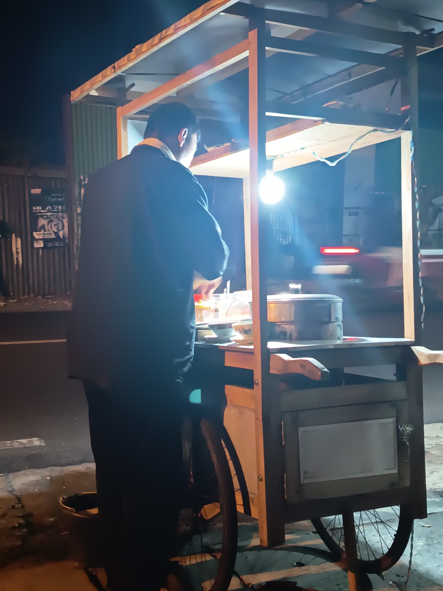 The image shows a street food cart at night, located on a street in Ngaglik, Indonesia. A person, likely the vendor, is standing behind the cart, facing away from the camera and towards the food preparation area. The cart is constructed from wood and has a simple roof supported by vertical beams. A bright incandescent bulb illuminates the workspace. On the cart's surface, there are containers of food and a steamer. In the background, the dark night sky is visible, along with a blurred view of a passing vehicle with red taillights, indicating it is on a road. To the left of the cart, a corrugated metal fence and some posters are visible. The ground appears to be asphalt and pavement, with some debris scattered around. The cart is supported by bicycle wheels at the rear.