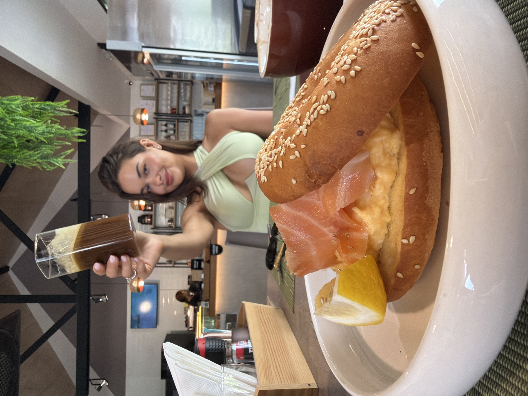 The image shows a woman in a restaurant in Choeng Thale, Thailand. She is holding a glass of a layered brown and yellow beverage. The restaurant appears to be modern with dark wood tones and a green plant. In the foreground is a bagel sandwich with salmon, egg, and a lemon wedge, plated in a white bowl. A wooden napkin holder containing white napkins is on the table, along with a salt shaker.