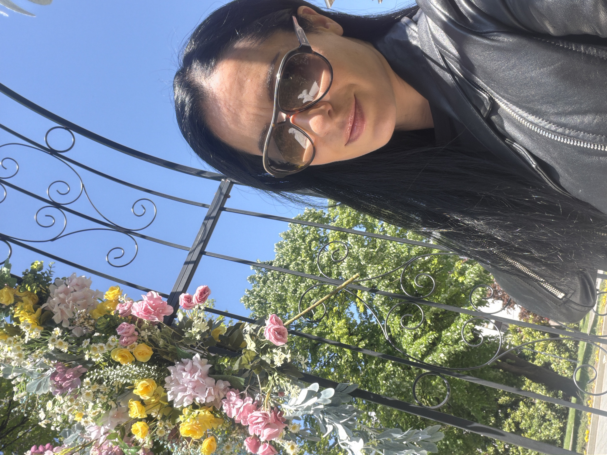 A woman wearing sunglasses and a black leather jacket poses for a selfie. The shot is taken at an angle, looking up at her against a clear blue sky. She is standing under a wrought-iron arch adorned with pink and yellow flowers. Behind the arch, green trees are visible. The location is Pleven, Bulgaria.