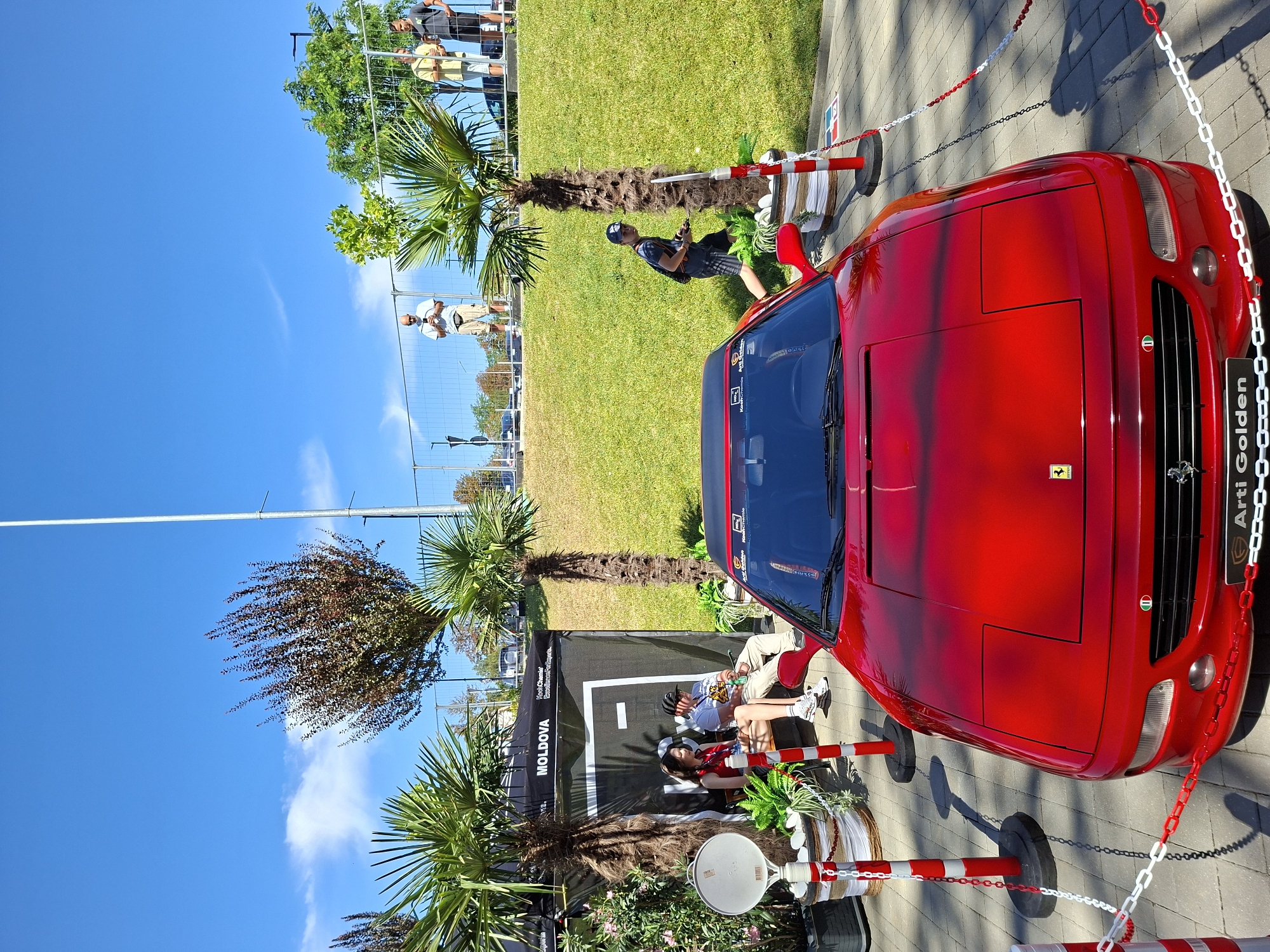 This image depicts an outdoor event in Stăuceni, Moldova, featuring a red Ferrari car as the central focus. The car is parked on a paved surface, cordoned off by red and white stanchions and chains. Two palm trees flank the car. In the background, there is a grassy hill with several people observing the scene. A black tent with "MOLDOVA" branding is visible behind the palm trees on the left, with two individuals sitting on a bench in front of it. A tall pole, possibly a flagpole, stands in the background. The sky is clear blue with scattered clouds.
