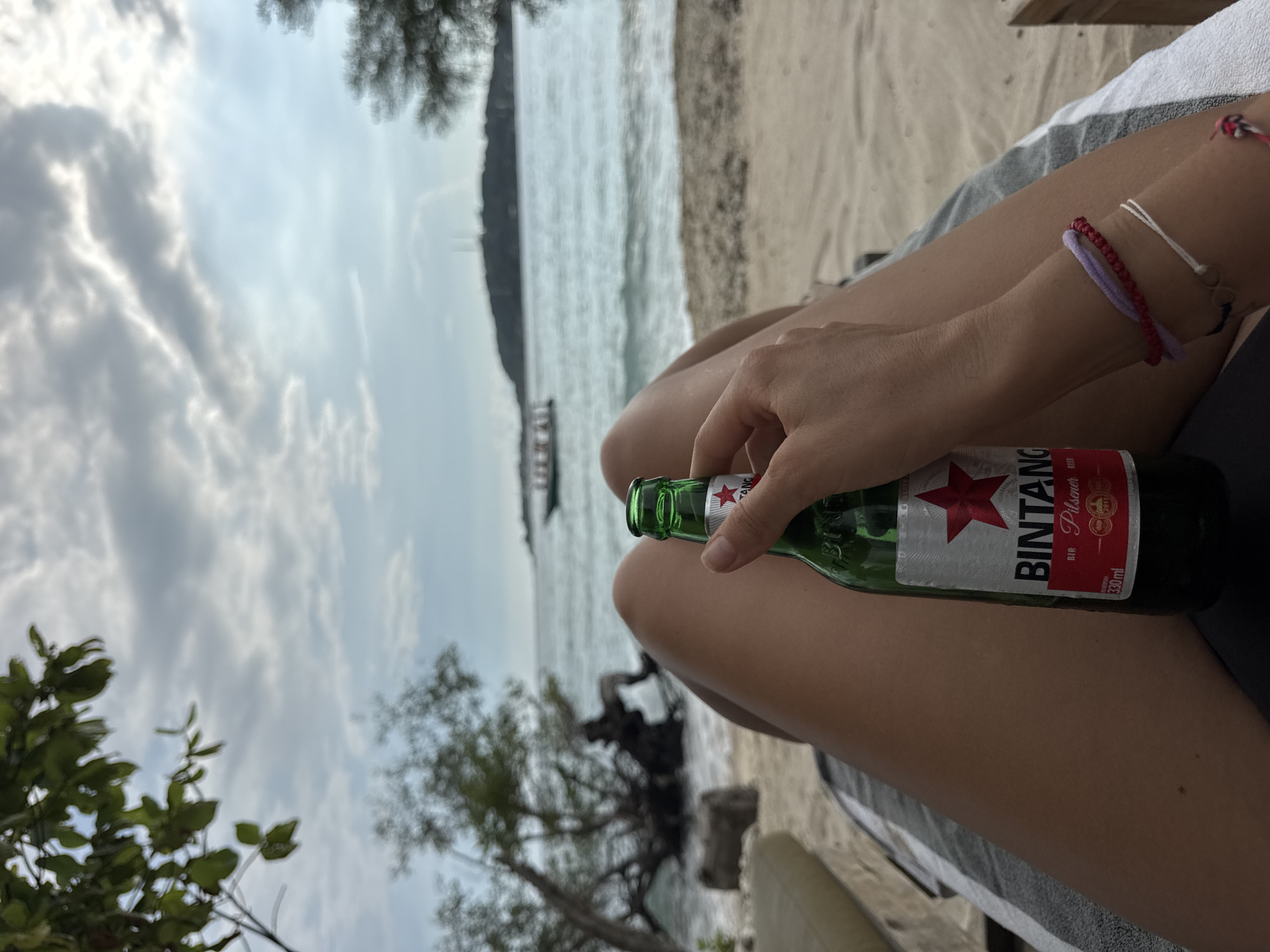 The image depicts a person holding a bottle of Bintang beer on a beach. The beach is located in Unknown, Indonesia. The person is sitting or reclining, as indicated by the positioning of the legs and the presence of a cushion or towel beneath. The beach has white sand. In the background, there is a view of the ocean and a silhouette of an island or landmass in the distance. Vegetation, possibly trees or shrubs, is visible in the foreground. The sky is cloudy. The person's wrist has a few bracelets.