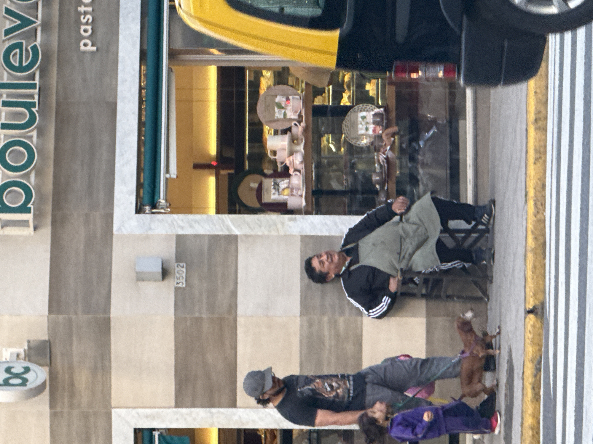 This image depicts an urban street scene in Buenos Aires, Argentina, featuring people, a dog, a commercial establishment, and a vehicle. The scene is set during the daytime under clear weather conditions.
In the foreground, a man in a black jacket with white stripes on the sleeves and a light-colored apron is seated on a black folding chair. He is looking towards the left side of the frame. Standing nearby is another man wearing a gray baseball cap, a dark t-shirt with a graphic design, and gray pants. He holds a leash attached to a small, brown dog that stands alertly next to him. A child or young person in a purple jacket is partially visible behind the standing man.
The background is dominated by the facade of a commercial building, possibly a shop or bakery, with alternating light and dark rectangular panels. Prominently displayed on the building is the text "boulevard pasteleria," indicating a pastry shop. A round sign with the letters "bc" is also visible. A street number, "3502," is affixed to the building. Through a large display window, various items, including cups and other merchandise, can be seen inside the establishment.
To the right of the people, a paved area with distinct white stripes and a yellow curb suggests a street or pedestrian crossing. The rear section of a yellow and black vehicle, possibly a taxi or delivery truck, is partially visible at the top of the frame. The overall activity captures a quiet moment of daily life in a city, with individuals and a pet pausing near a local business.