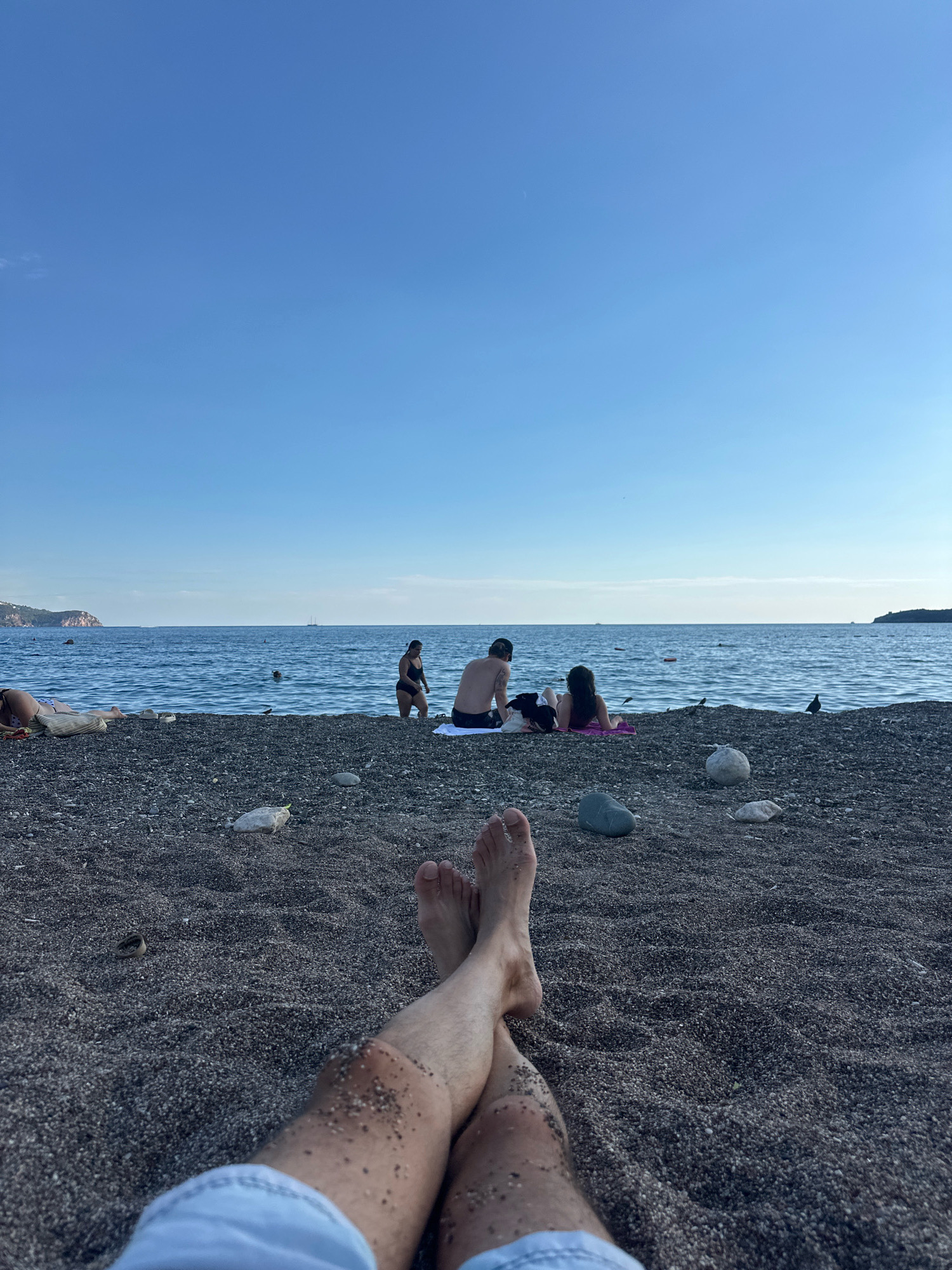 The image depicts a beach scene in Boreti, Montenegro, under a clear blue sky. The foreground is dominated by the legs and feet of a person lying on the dark, pebbly beach, with sand clinging to their skin. In the mid-ground, several individuals are relaxing on the shore. Two people, a man and a woman, are seated on a towel, facing the sea. Further to the left, another person is lying on a towel. A woman in a dark bikini is walking along the water's edge. The sea stretches out to the horizon, where distant landmasses and a few small boats are visible. The beach is sparsely populated, with a few scattered pebbles and some pieces of litter. A bird is perched on the sand near the water. The overall atmosphere is one of relaxation and enjoyment of a sunny day at the beach.
