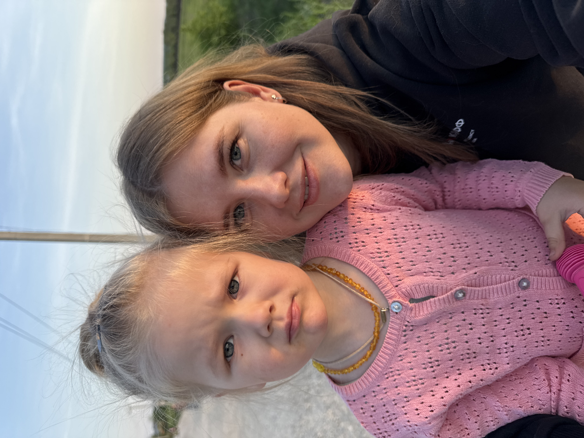 The image depicts a selfie of a young woman with light brown hair and a young girl with blonde hair. They are closely positioned, with the woman on top and the girl below. The woman is wearing a black hoodie. The girl is wearing a pink, knitted cardigan and a yellow bead necklace. The background shows a blurry landscape with hints of greenery and a sky. The location is Горный, Russia.
