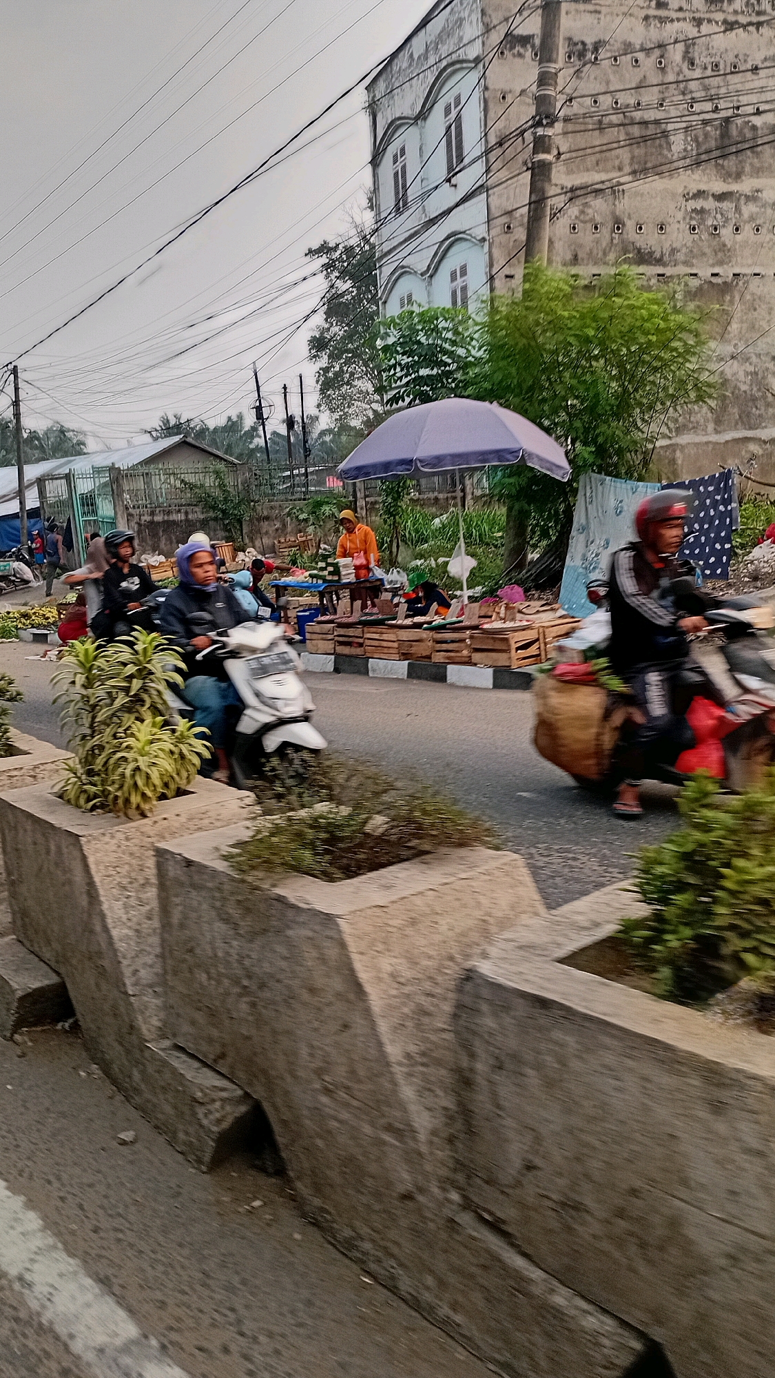 The image depicts a street scene in Jambi, Indonesia, possibly during daylight hours under an overcast sky. The foreground features a concrete planter box with greenery. The midground shows a paved road with several people on motorcycles and market stalls lining the side.
On the left, a white scooter with two individuals, one driving and one a passenger wearing a blue headscarf, moves away from the camera. To the right of the road, a market is set up with wooden crates displaying various goods, and a person in an orange shirt stands behind a table. A purple umbrella provides shade over a section of the market.
Further to the right, a person wearing a helmet on a red and black motorcycle is seen moving towards the camera, carrying a large woven basket on the back. The background includes a multi-story building with distinct architectural features, electrical wires, trees, and other smaller structures suggestive of a residential or commercial area. The overall impression is of a busy street with roadside commerce.