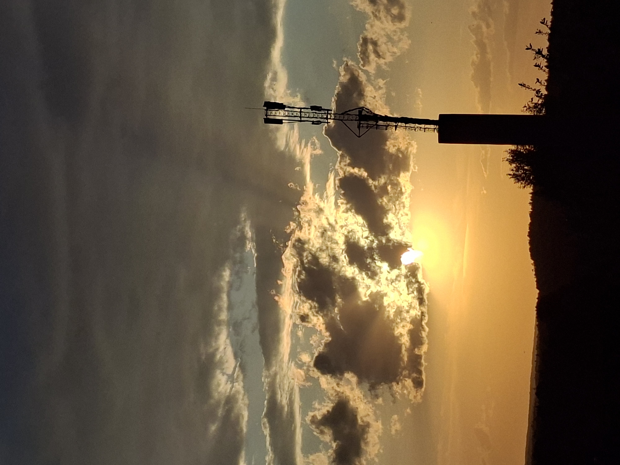 The image depicts a sunset with the sun partially obscured by clouds. In the foreground, silhouetted against the bright sky, is a tall structure that appears to be a communication tower or a similar type of structure, with a cylindrical base and antennae extending from it. To the right of the tower, the edge of a dark, possibly vegetated, area is visible, forming a silhouette against the lighter sky. The sky itself displays a gradient of colors, ranging from a dark, possibly stormy gray on the left to a bright, golden-yellow hue around the setting sun. The clouds exhibit varied textures and are illuminated by the sun, creating a dramatic effect. The location is Karsha, Russia.