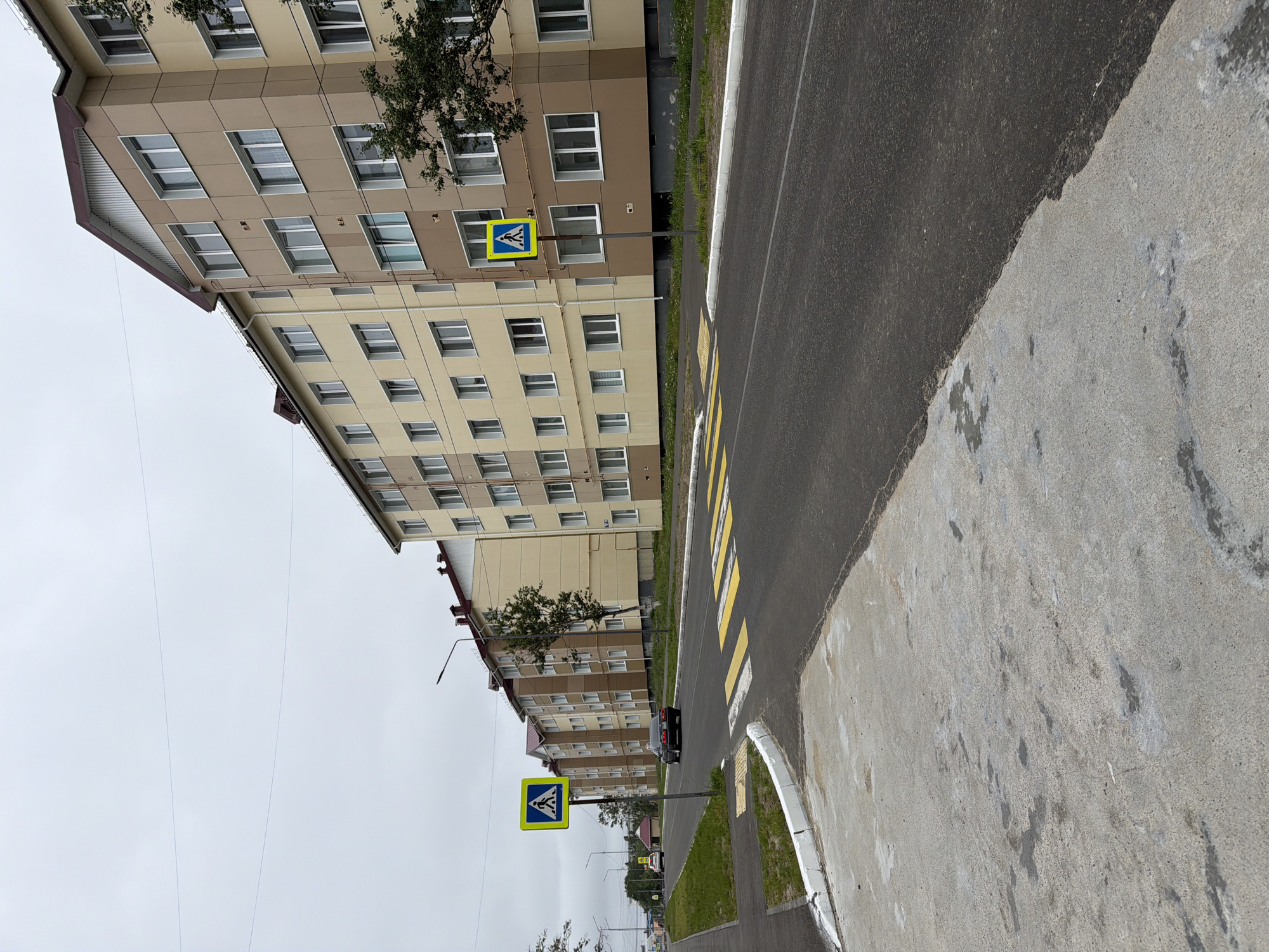 The image shows a street in Okha, Russia, with a row of multi-story apartment buildings on the right side of the road. The buildings are light yellow and brown with red roofs and numerous windows. A pedestrian crossing is marked with yellow and white stripes on the asphalt road. A blue and yellow pedestrian crossing sign is visible on the left side of the road. A dark-colored car is driving on the road in the middle distance, and another white car is visible further down the road. The sky is overcast. The foreground shows a concrete sidewalk adjacent to the road.
