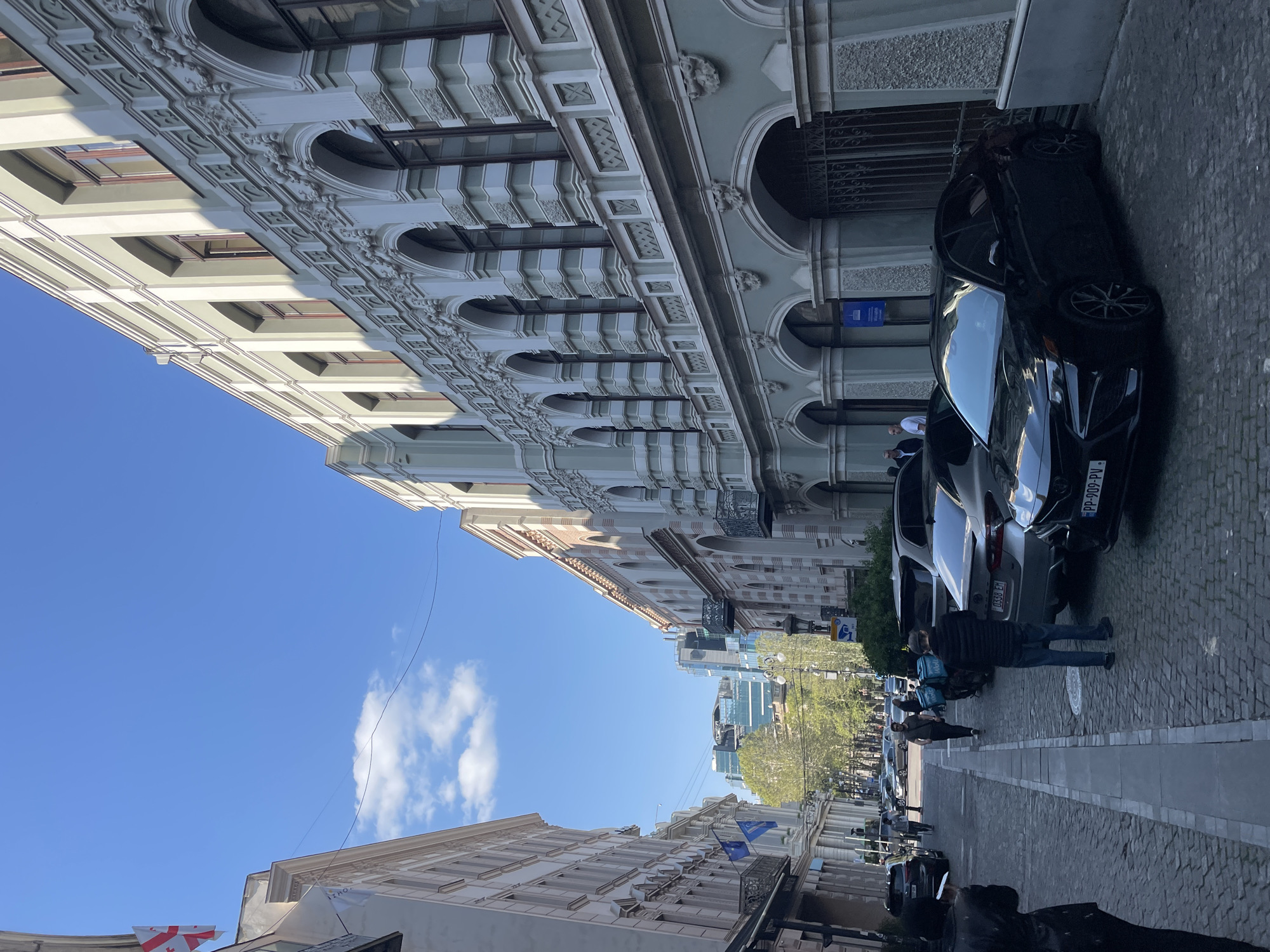 The image displays a bustling street scene in Tbilisi, Georgia, under a clear blue sky with scattered white clouds. The perspective is looking down a cobblestone street lined with buildings that showcase a mix of architectural styles.
On the left, a large, ornate building with intricate classical architecture dominates the foreground. It features multiple stories with arched windows, decorative moldings, and cornices, with parts illuminated by sunlight. Further along this side, other buildings are visible, one adorned with a white Georgian flag displaying five red crosses, and another with a small white flag bearing the text "IZHO".
On the right side of the street, several cars are parked on the cobblestones. A prominent black car in the mid-ground displays a Georgian license plate, "PP-009-PV". Behind it, a silver car is partially visible, showing "GB-0350 BM" on its license plate. People are seen walking on the street and sidewalk. In the mid-ground, a person wearing a dark jacket and blue jeans stands near the parked cars. Further down the street, a delivery person is identifiable by two light blue square delivery bags carried on their back. Blue rectangular signs are attached to the buildings on the right.
The background reveals a blend of old and new, with modern glass high-rise buildings and lush green trees suggesting a contemporary urban extension beyond the historic street. The scene depicts everyday activity on a sunny day in the city.